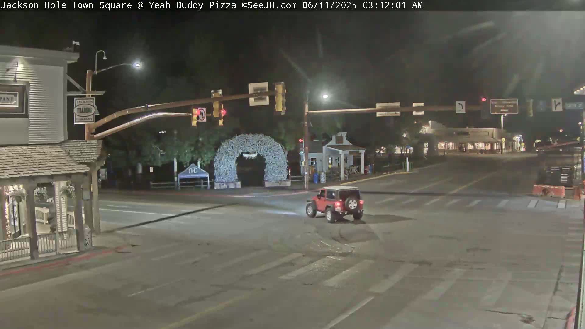 A red Jeep drives through a mostly empty town square at night, with an archway decorated with white branches in the background.