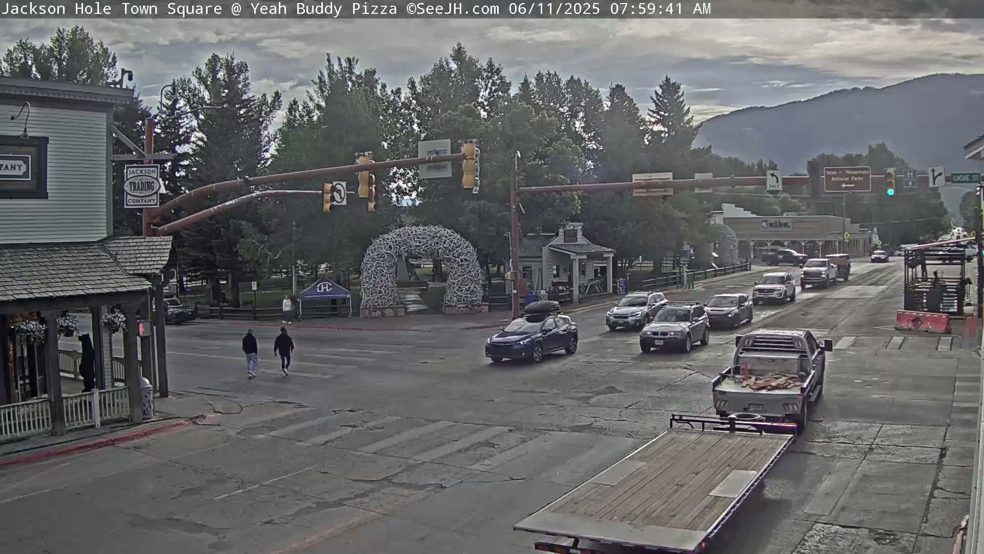 A town square with overcast skies, several cars at an intersection, and two pedestrians crossing the street, with a large antler arch in the background.
