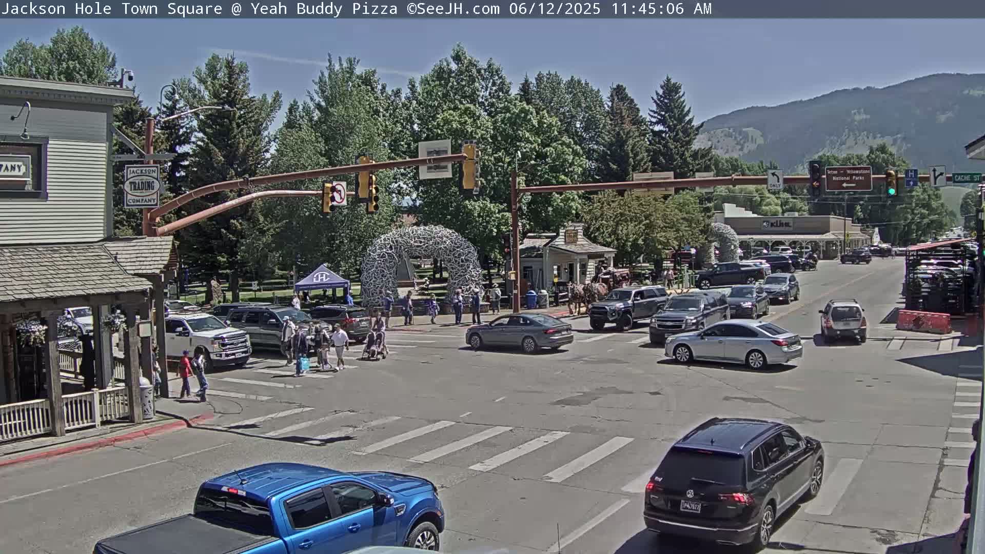 A sunny town square intersection with pedestrians and numerous vehicles, some stopped at a crosswalk, under a clear sky and surrounded by trees and buildings.