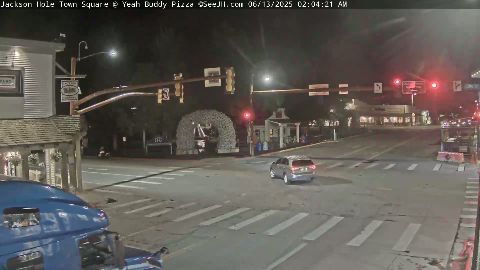 A nighttime view of a town square intersection with a silver SUV driving through, under streetlights, with a partially visible blue truck in the foreground.