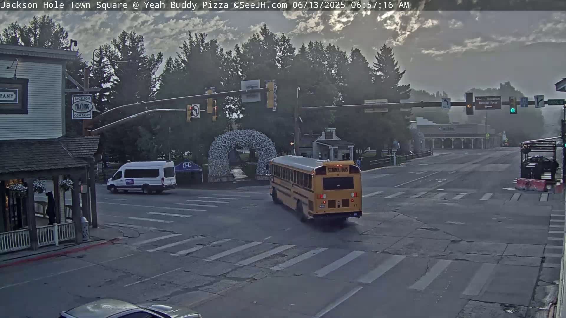 A mostly overcast morning shows a school bus and a white van at a town square intersection with pedestrian crosswalks, under a hazy sky.