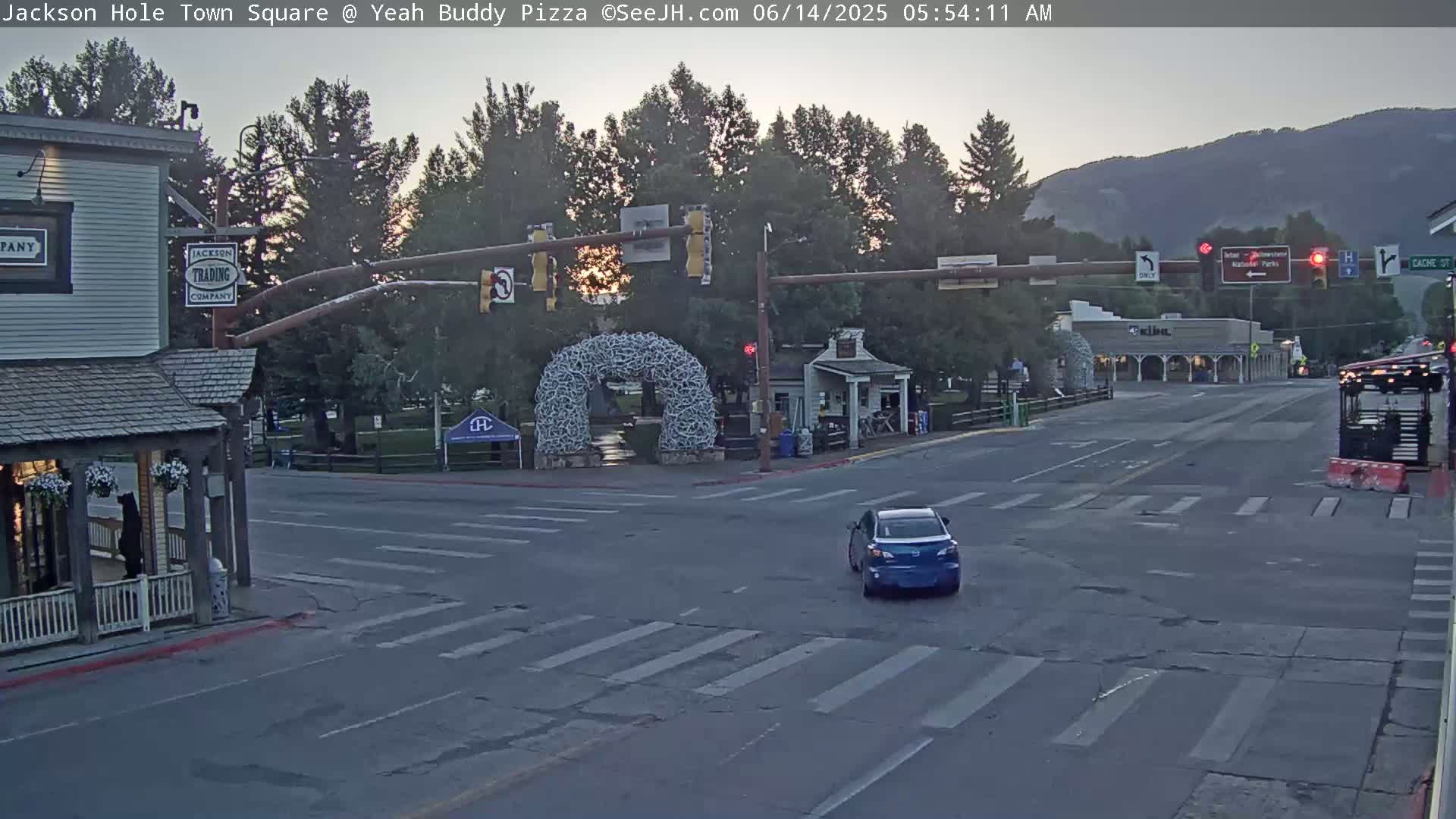 A mostly empty town square at sunrise, with a blue car at an intersection, under a partly cloudy sky.
