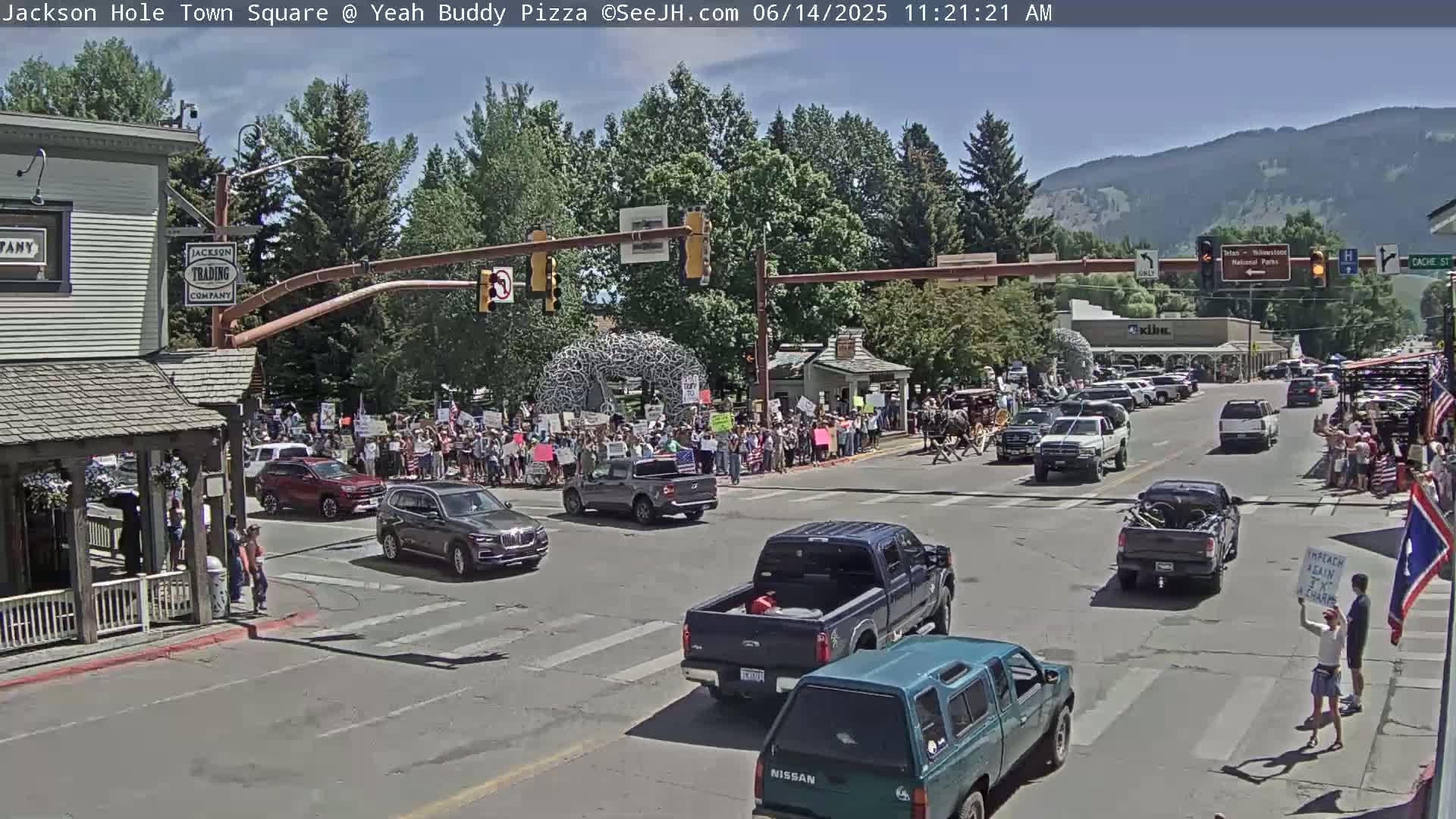 A large group of people are gathered in a town square on a sunny day, with several vehicles stopped at a crossroads.