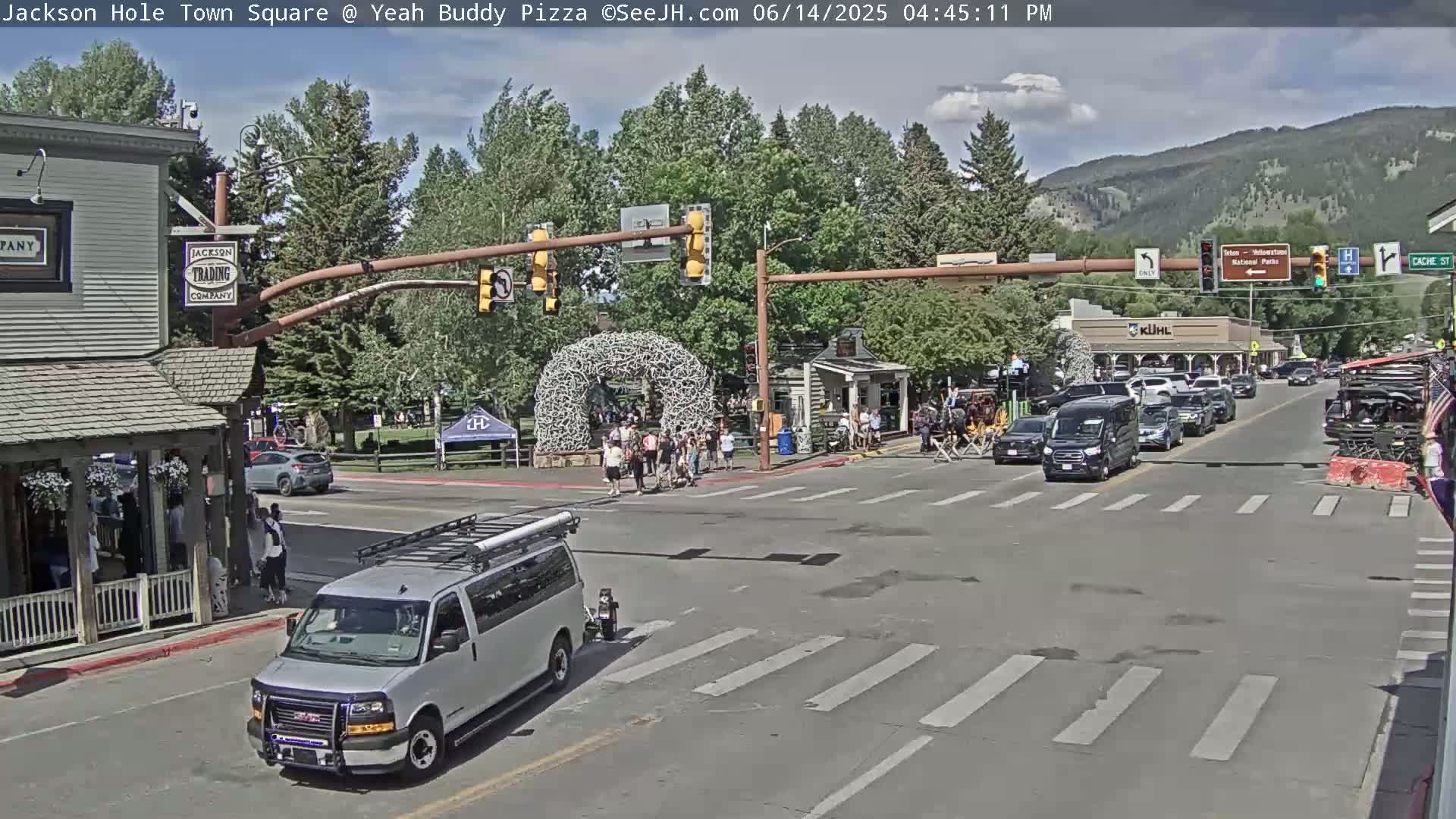 A large group of people are gathered in a town square on a sunny day, with several vehicles stopped at a crossroads.