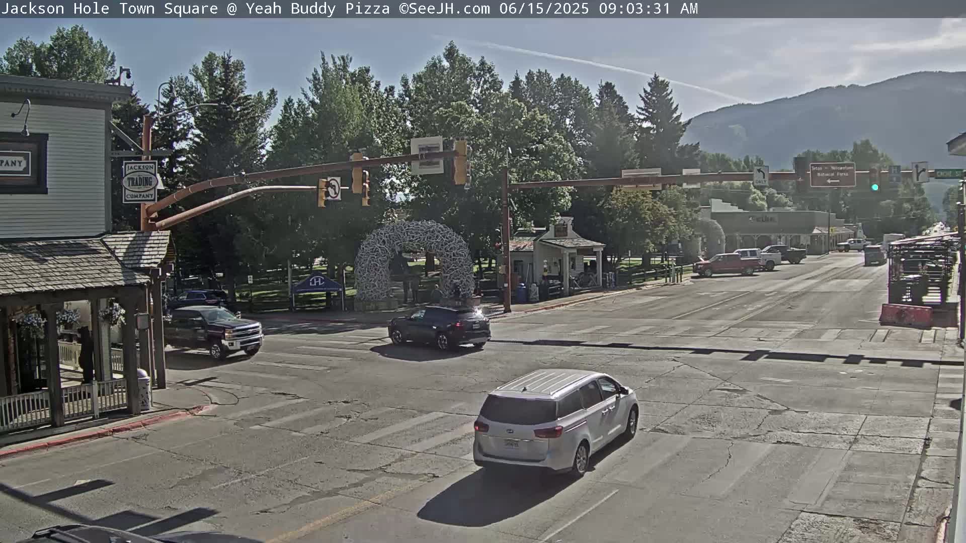 A sunny town square scene shows a street intersection with several vehicles, including a minivan, and buildings with trees in the background, under a clear sky.