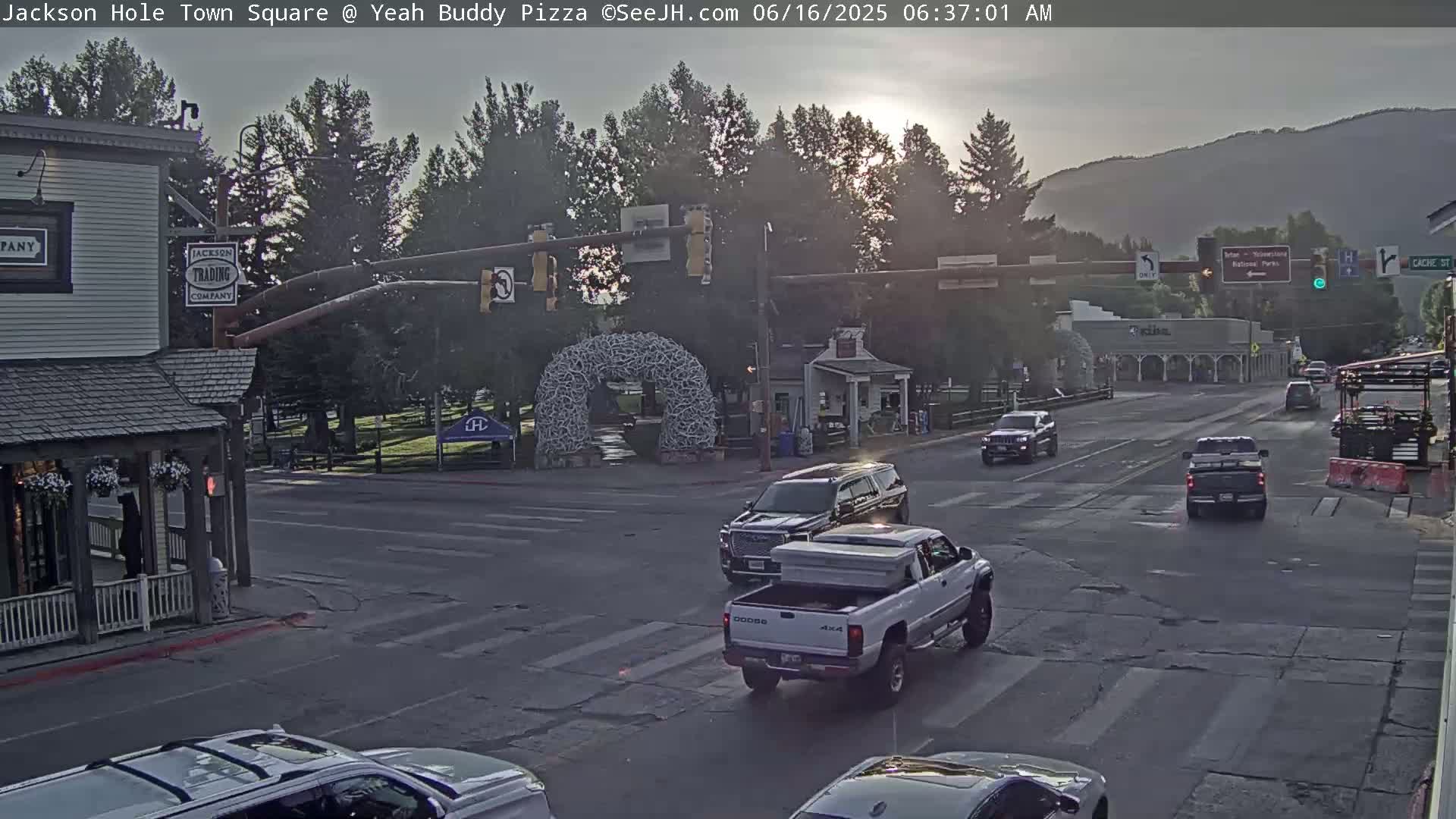 A mostly cloudy sunrise illuminates a town square with several vehicles at an intersection,  and buildings with some greenery visible in the background.