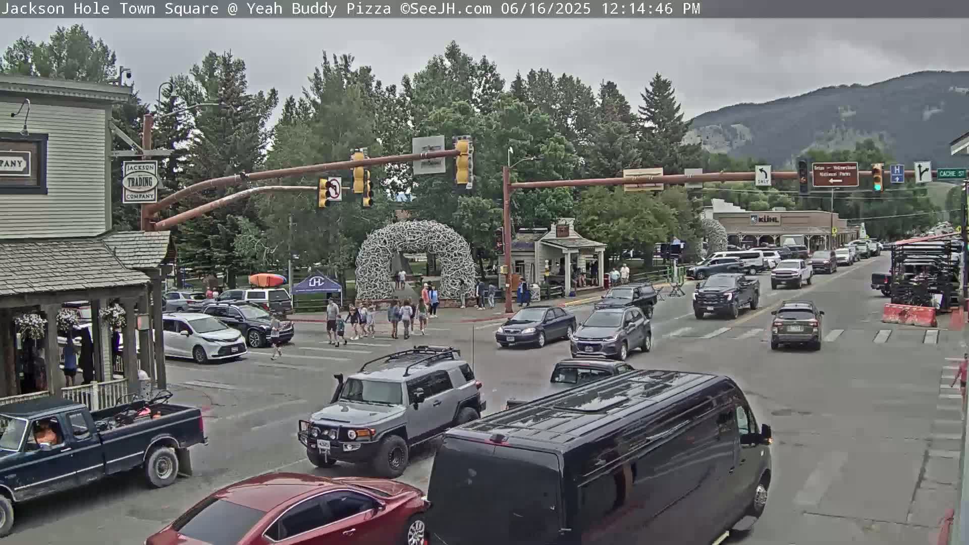 A town square with pedestrians and numerous vehicles at an intersection under an overcast sky, near a large antler arch.