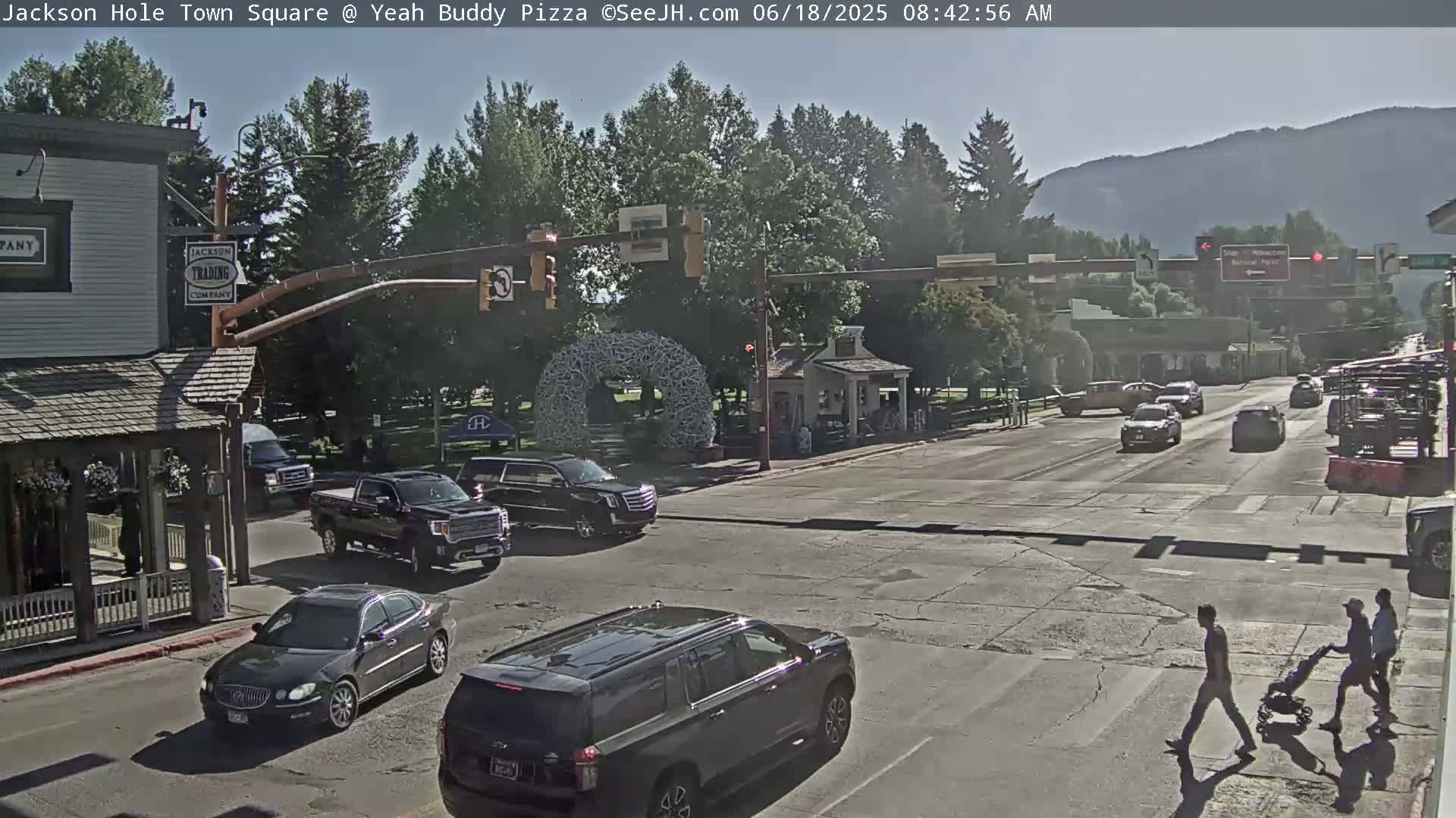 A sunny street scene shows a town square with several vehicles at an intersection, pedestrians crossing, and a large antler-like sculpture in the background.