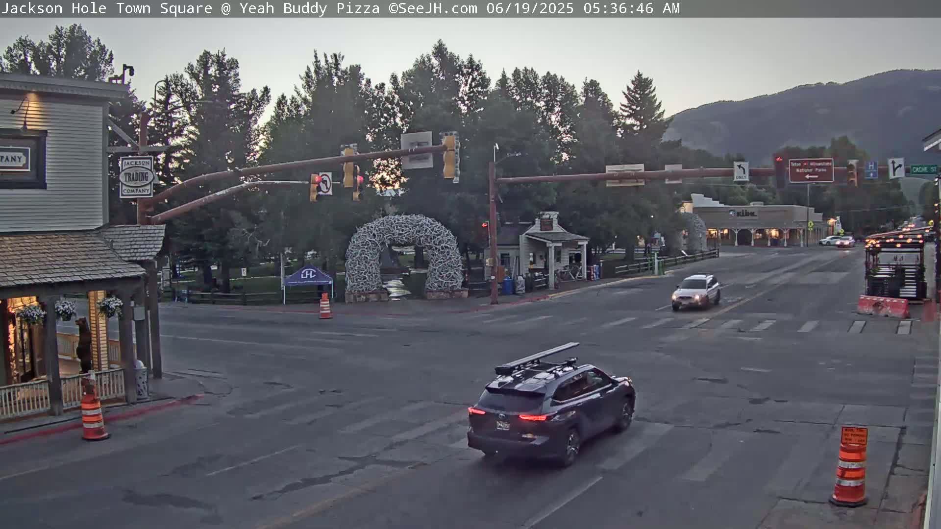 A mostly empty town square at dusk, with a few cars driving by, under a mostly clear sky.