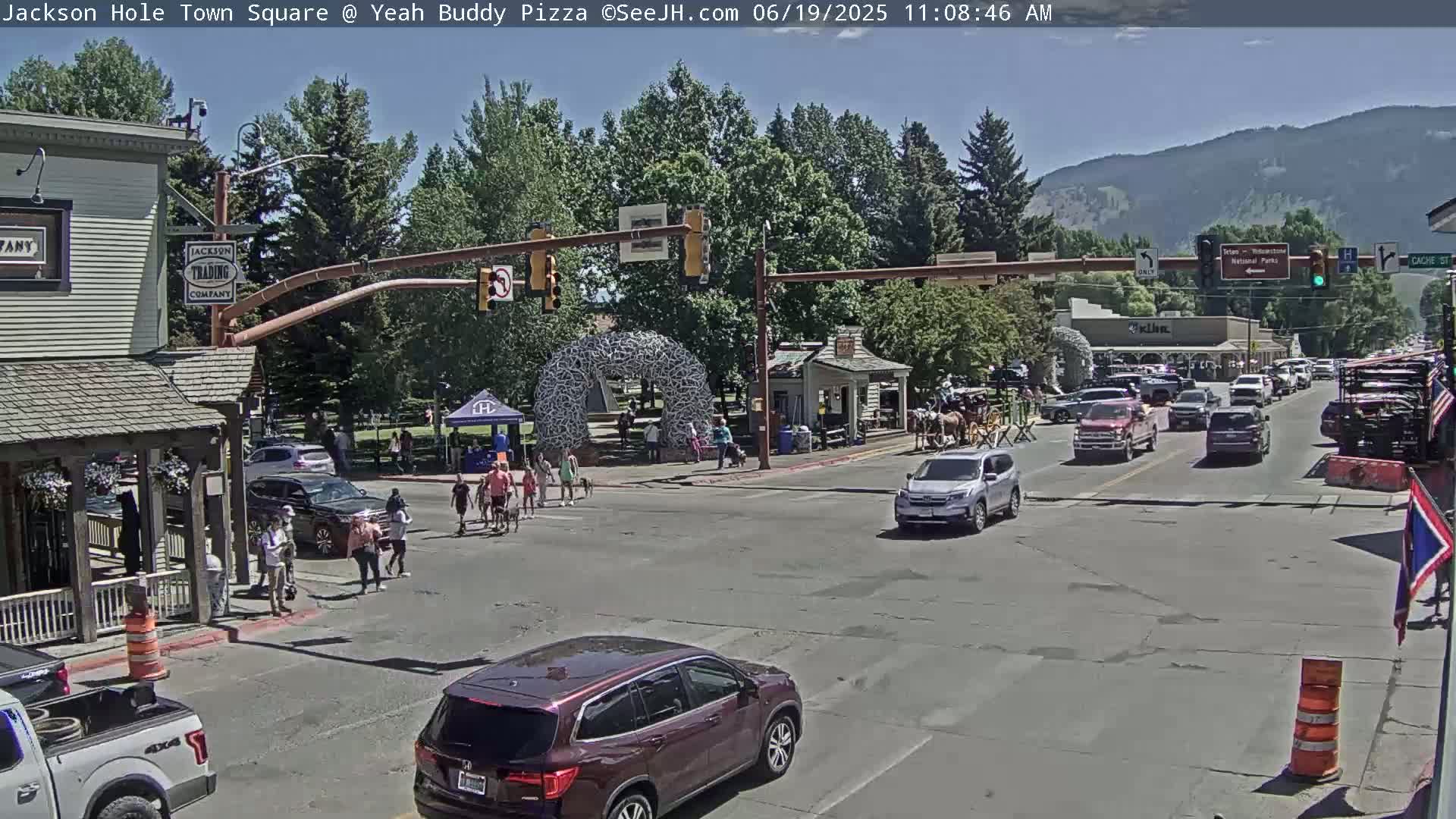 A sunny town square scene shows pedestrians crossing a street with several vehicles, including an SUV and a pickup truck, while a horse-drawn carriage is visible in the background.