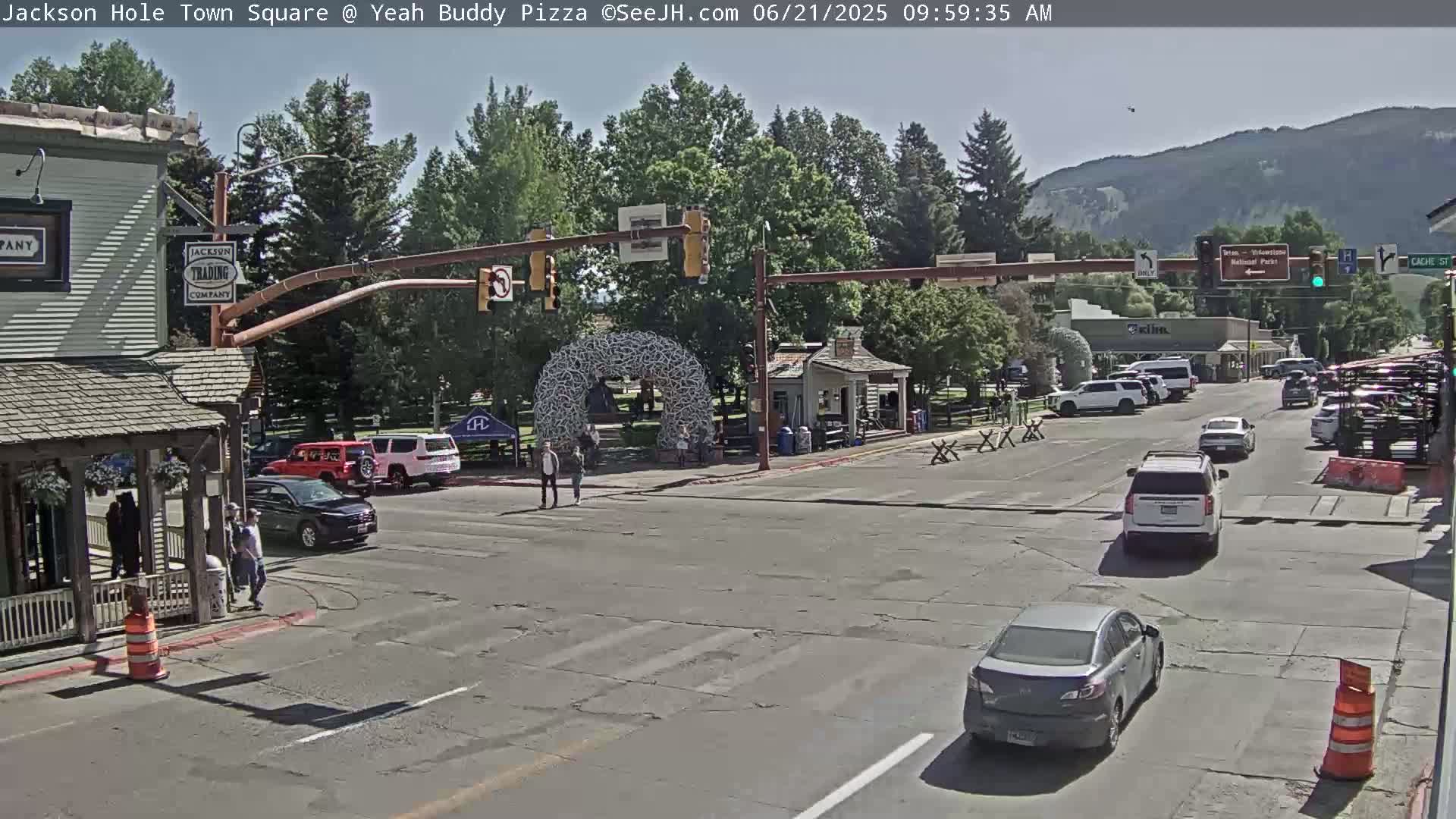 A sunny town square scene shows several cars at a crosswalk, pedestrians walking near buildings, and mountains in the background.