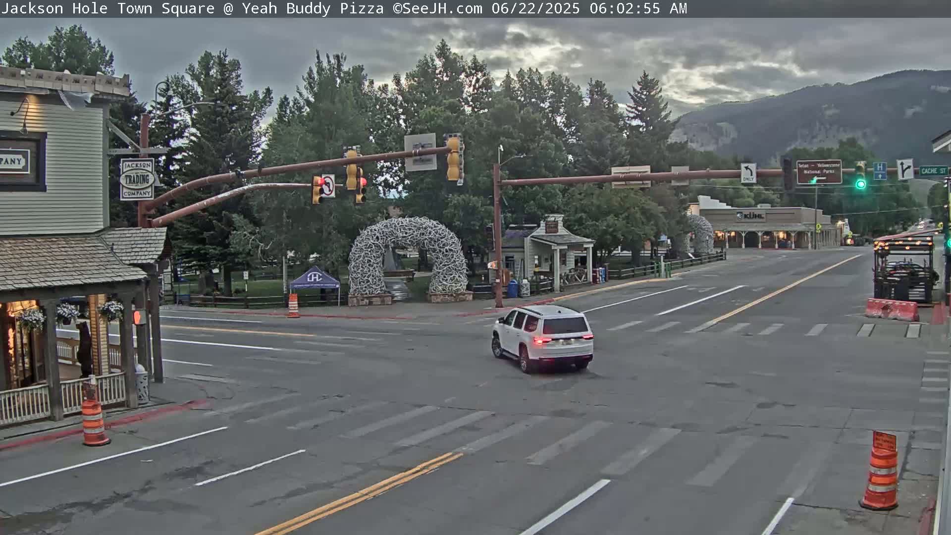 A white SUV drives through a town square intersection under an overcast sky, with mountains in the background and buildings lining the streets.