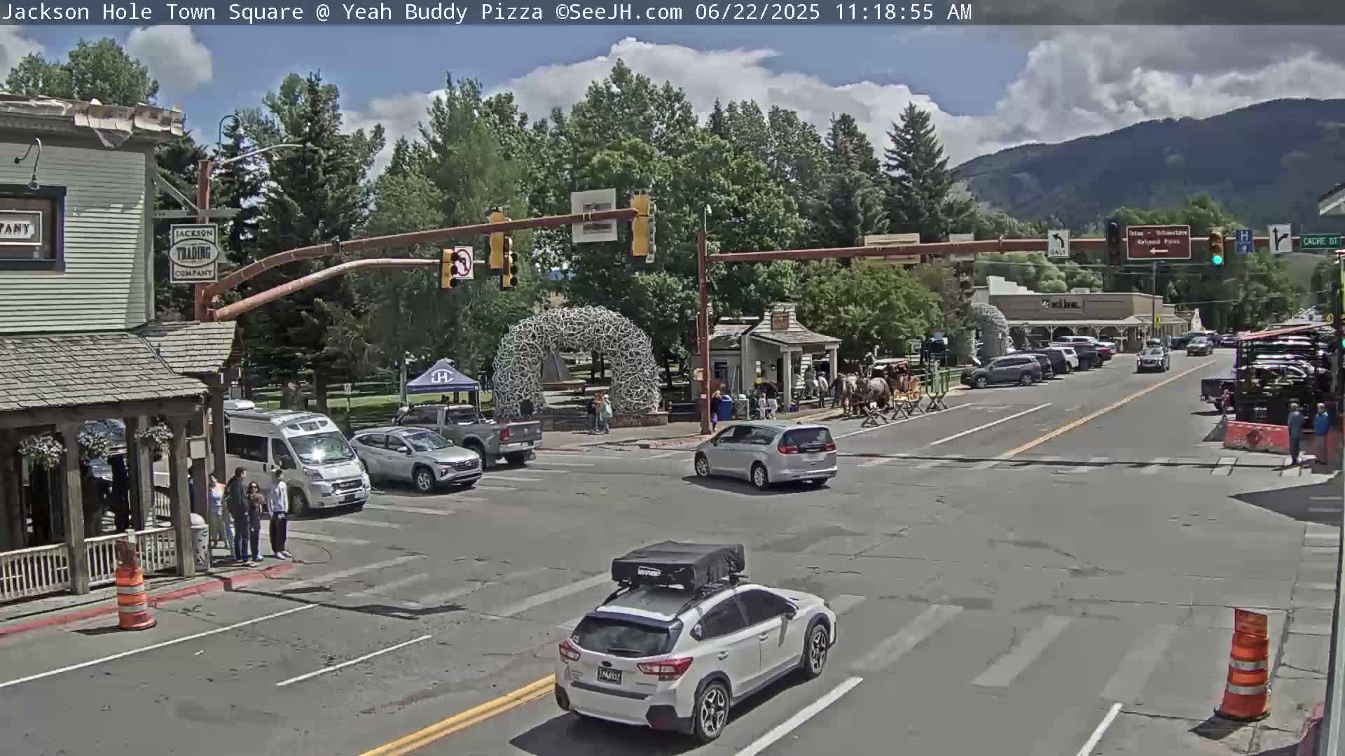A partly cloudy day in a town square shows several vehicles at an intersection, pedestrians on a sidewalk, and buildings along the street, with mountains visible in the background.