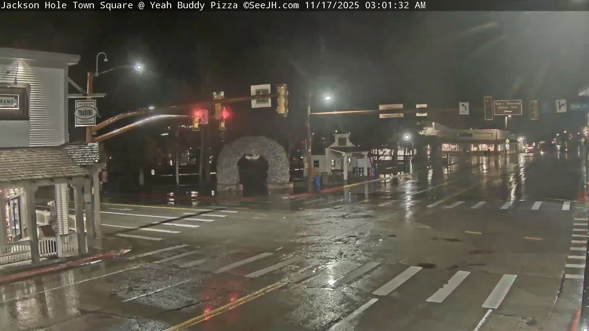 A wet street intersection at night is illuminated by streetlights, featuring an antler archway in the middle, various commercial buildings with lights, and reflections on the pavement from recent rain.