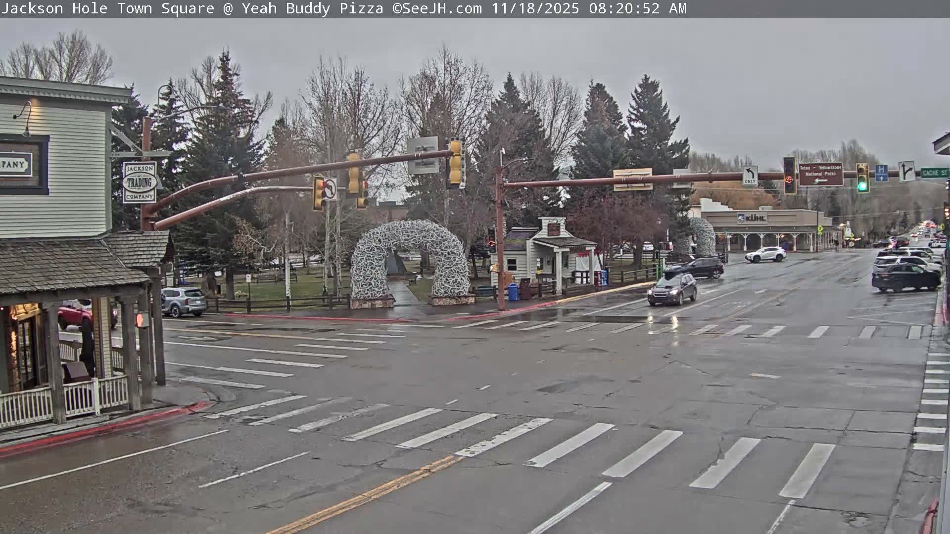 A wet street intersection at night is illuminated by streetlights, featuring an antler archway in the middle, various commercial buildings with lights, and reflections on the pavement from recent rain.