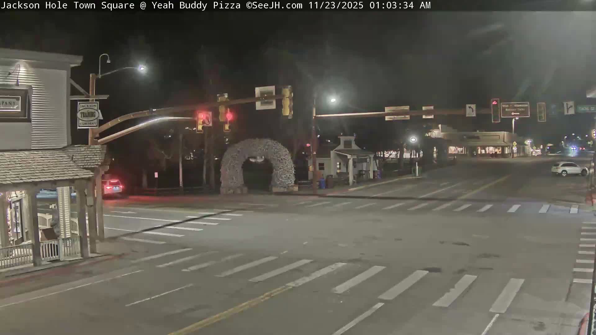A quiet town square intersection is depicted on a clear, dark night, featuring illuminated buildings, streetlights, an archway made of branches, and a few cars on the street.