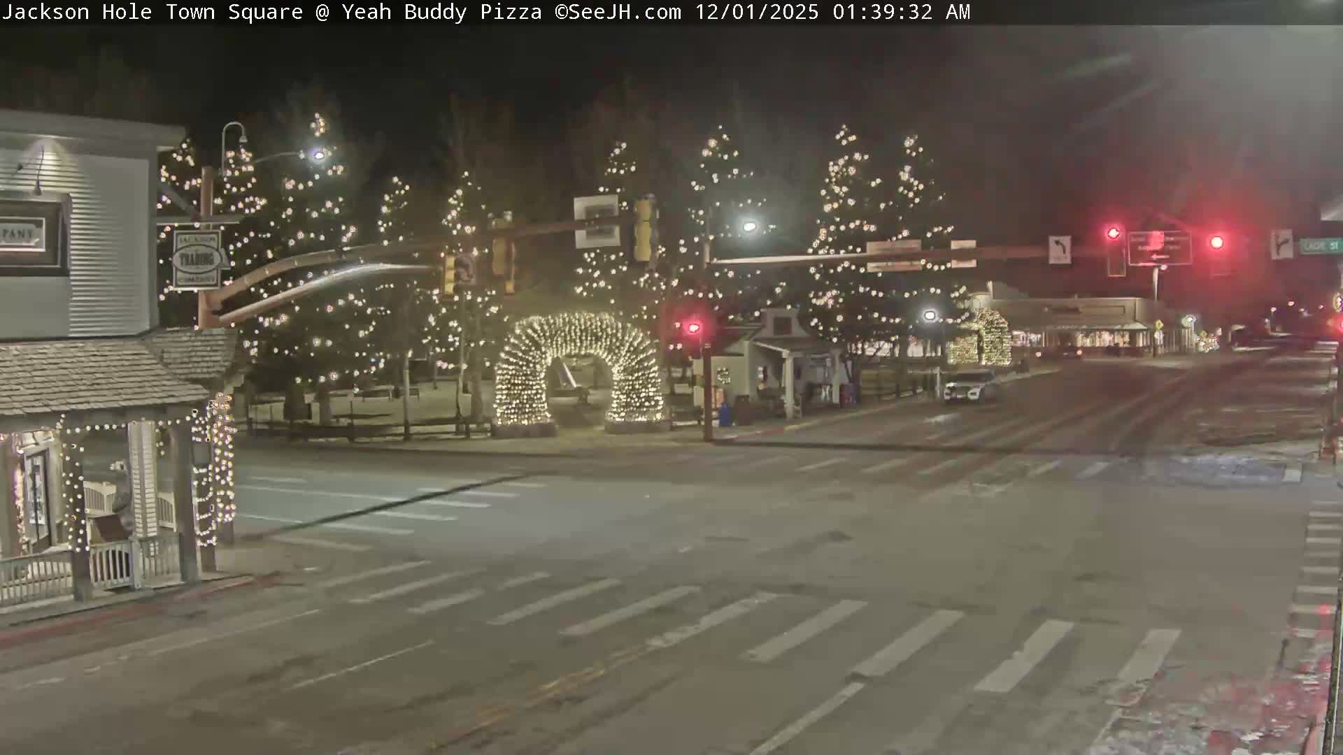 A town square is brightly illuminated at night with numerous white lights adorning trees, buildings, and a prominent archway, under clear weather conditions.