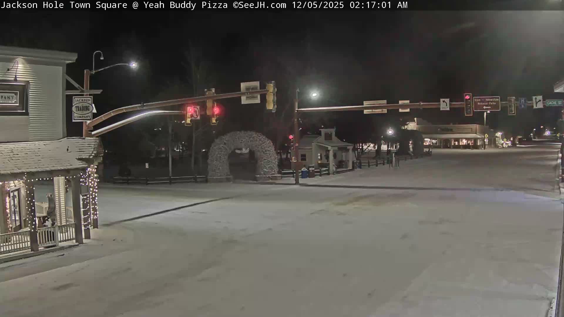 A quiet, snow-covered town square is illuminated at night by streetlights and building lights, with clear and cold weather conditions, showing traffic signals and a distinctive antler archway.