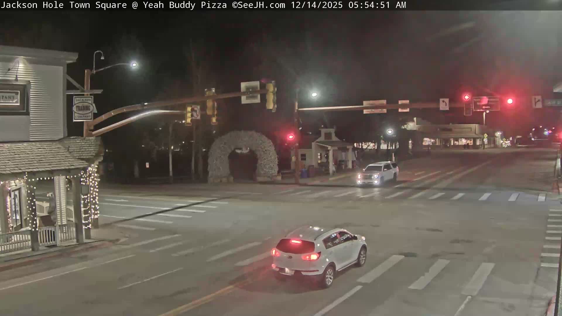 A quiet, snow-covered town square is illuminated at night by streetlights and building lights, with clear and cold weather conditions, showing traffic signals and a distinctive antler archway.