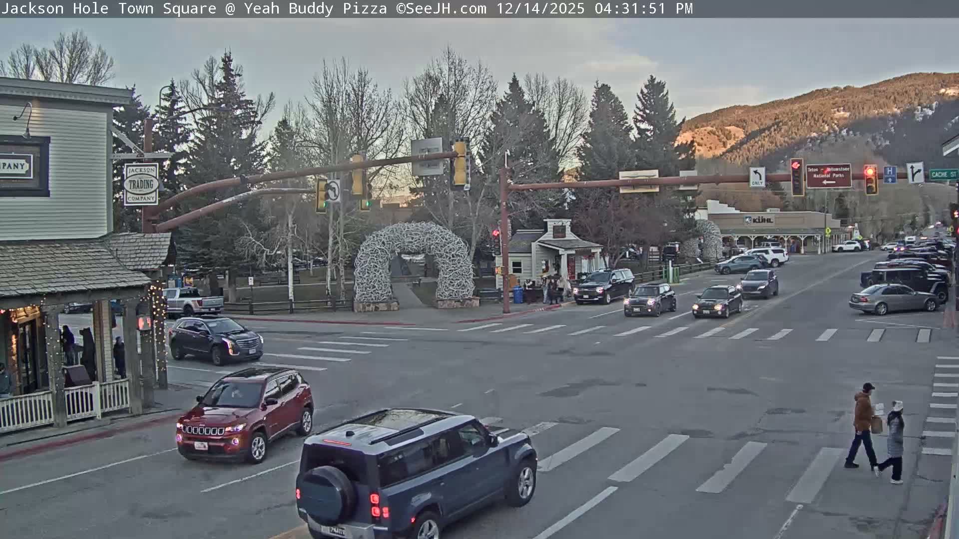 A quiet, snow-covered town square is illuminated at night by streetlights and building lights, with clear and cold weather conditions, showing traffic signals and a distinctive antler archway.