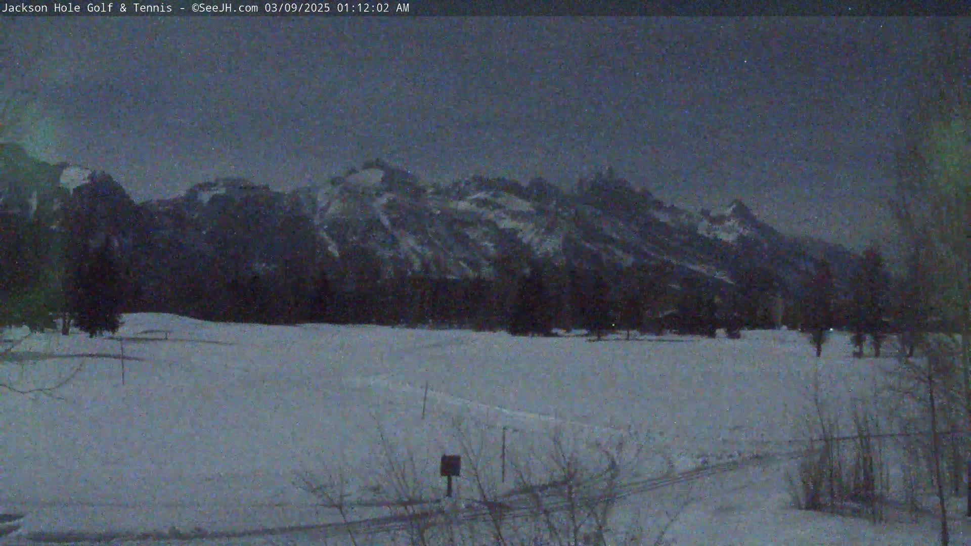 A snow-covered field stretches out before a dark, snow-capped mountain range under a twilight sky.