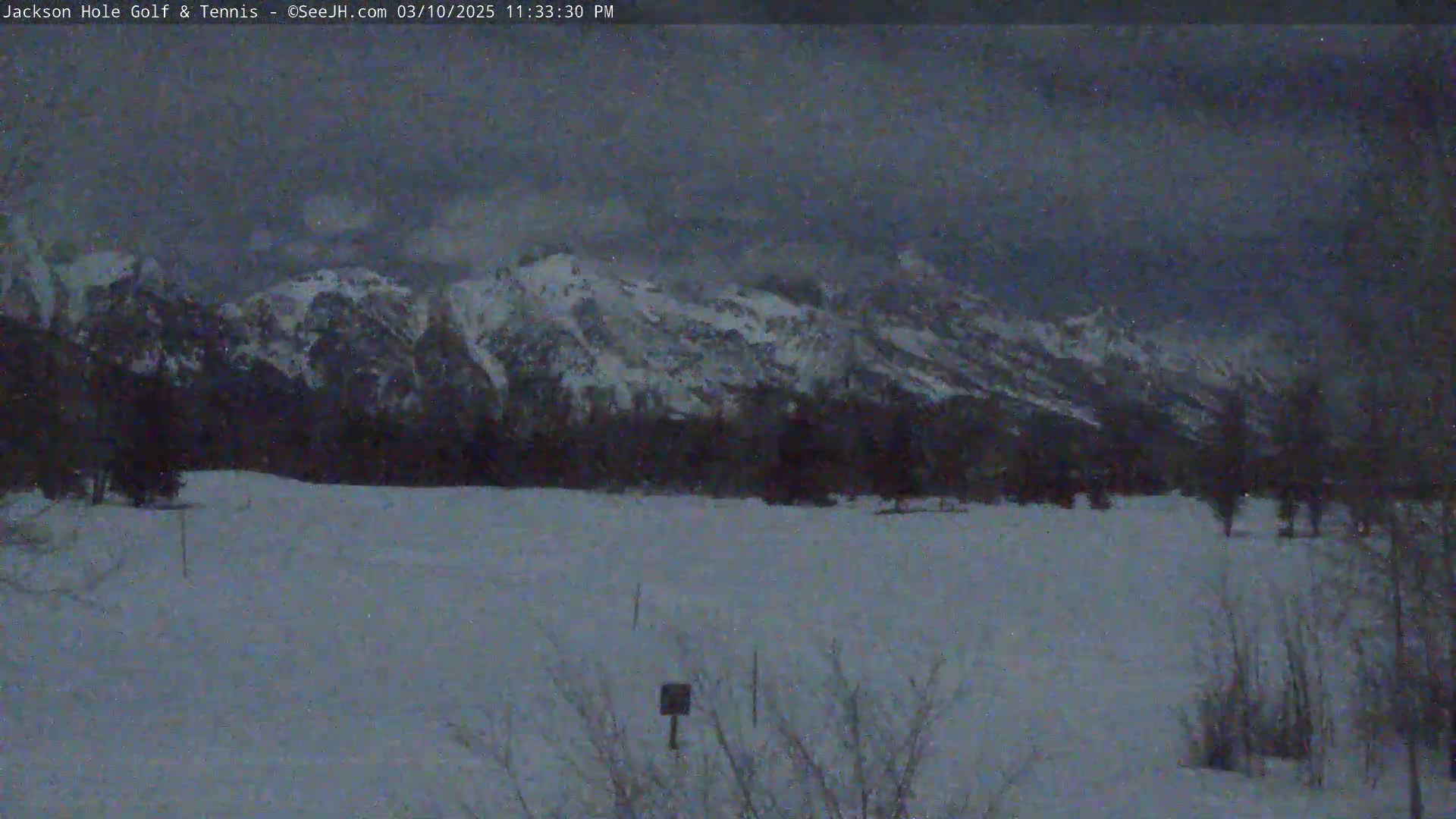 A snow-covered field stretches out before snow-capped mountains under a dark, overcast sky.