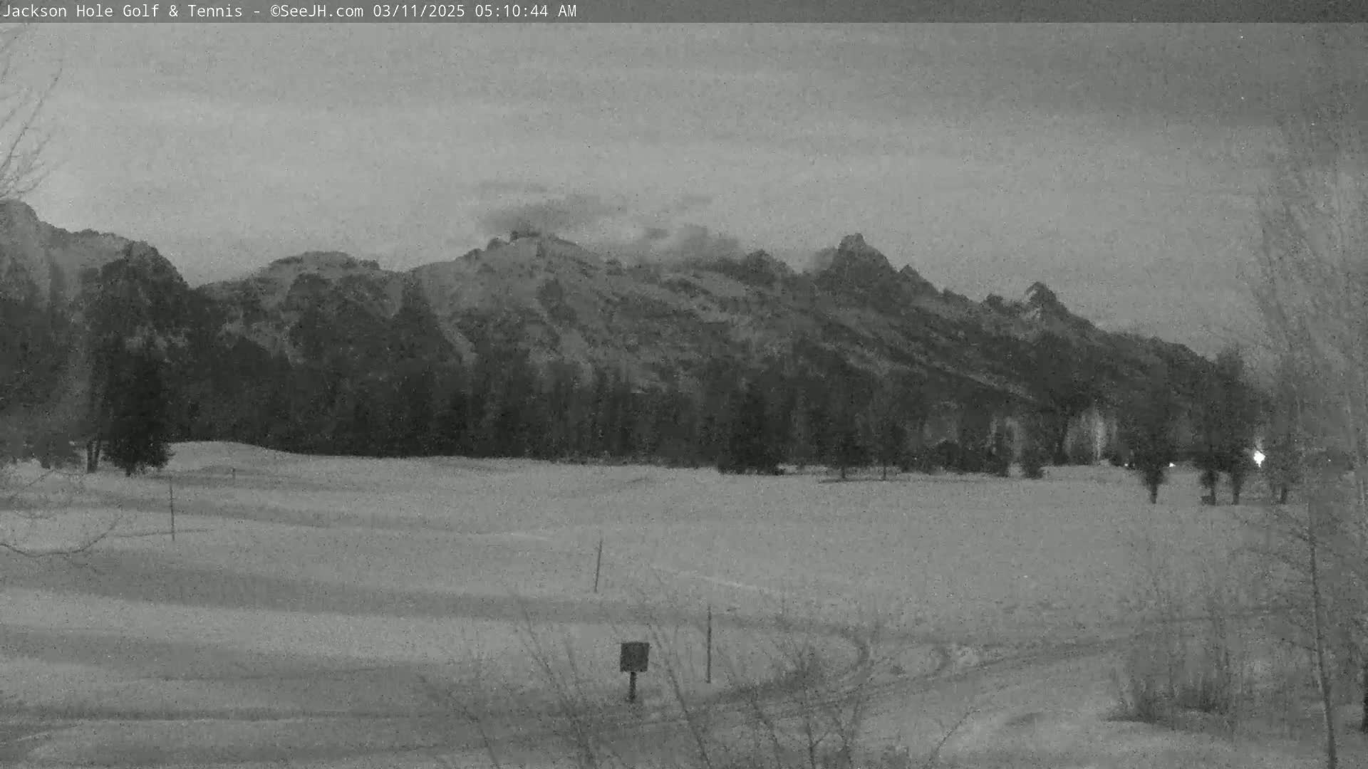 A snow-covered field stretches out before a range of dark mountains under a cloudy, overcast sky.