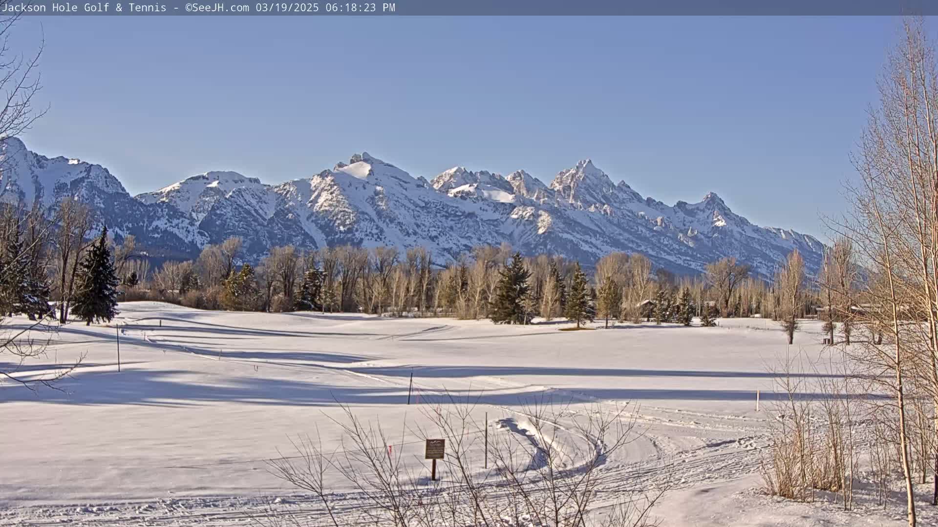 A snow-covered golf course with leafless trees and a snow-capped mountain range in the background under a clear, sunny sky.