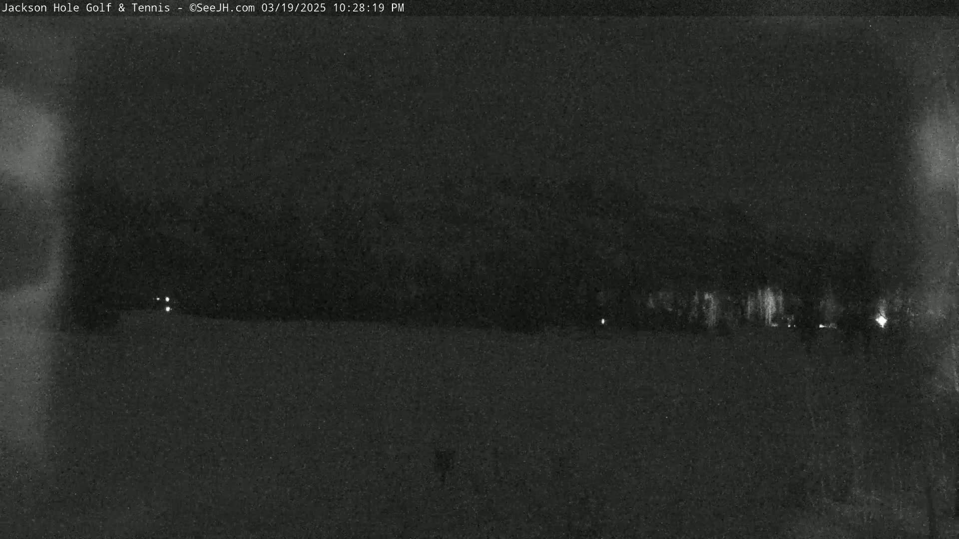 A nighttime, low-light view of a flat, dark landscape with a few faint lights in the distance.
