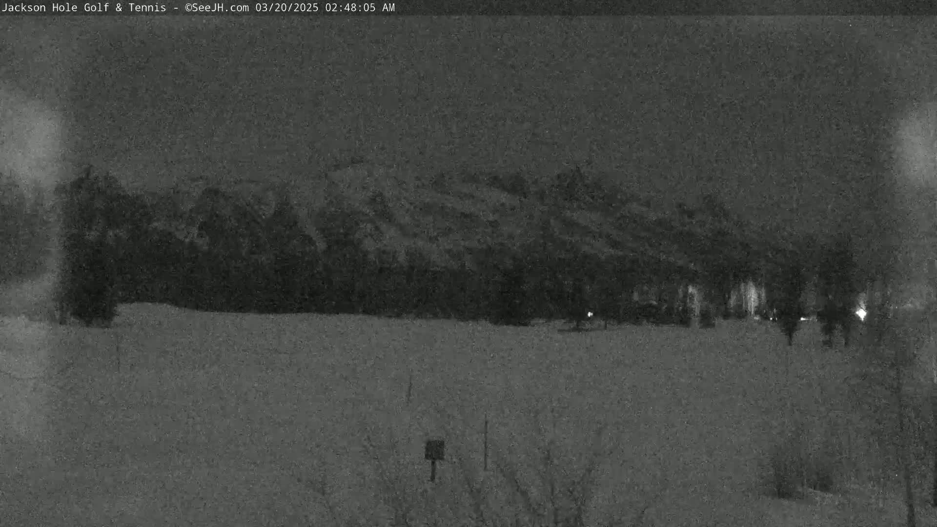 A nighttime, low-light view shows a snow-covered field in front of a dark, snow-dusted hillside under a cloudy sky.
