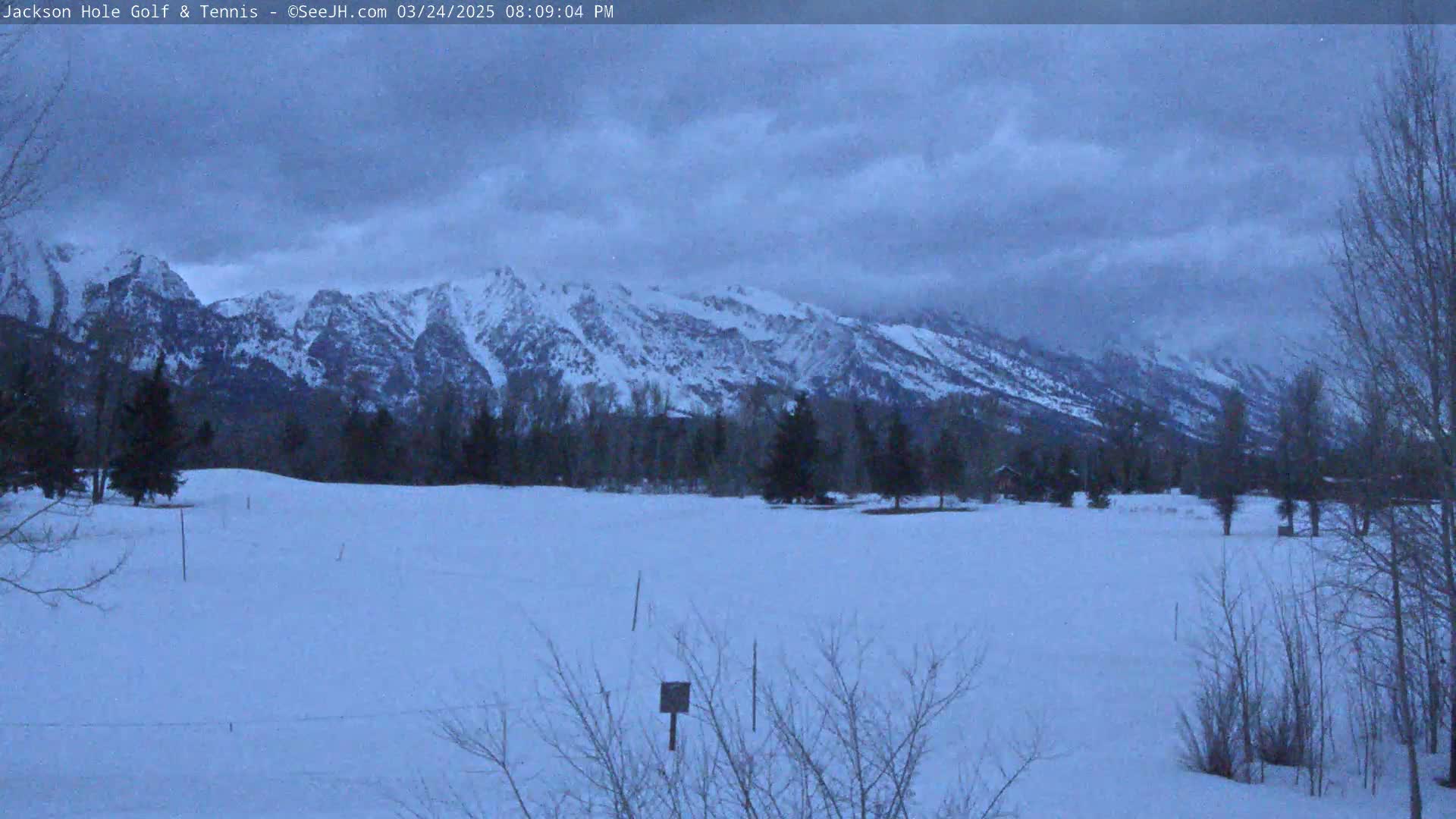 A snow-covered field stretches out before a range of snow-capped mountains under a cloudy, dusky sky.