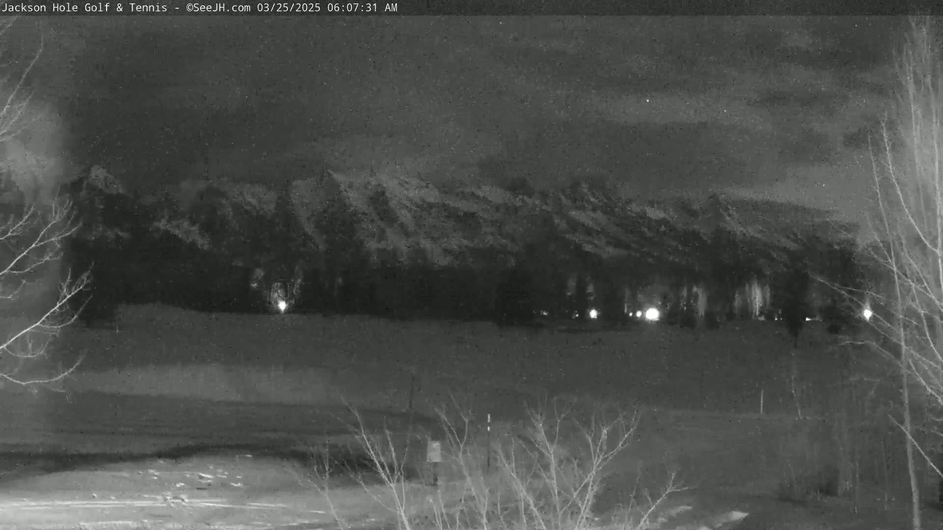 A nighttime, snow-covered mountain range is visible in the distance, with bare trees and a few distant lights in the foreground under a cloudy sky.
