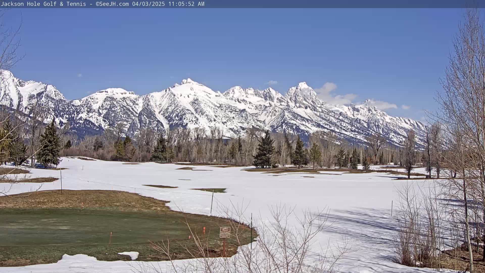 A partially snow-covered golf course lies in the foreground with a backdrop of snow-capped mountains under a clear blue sky.