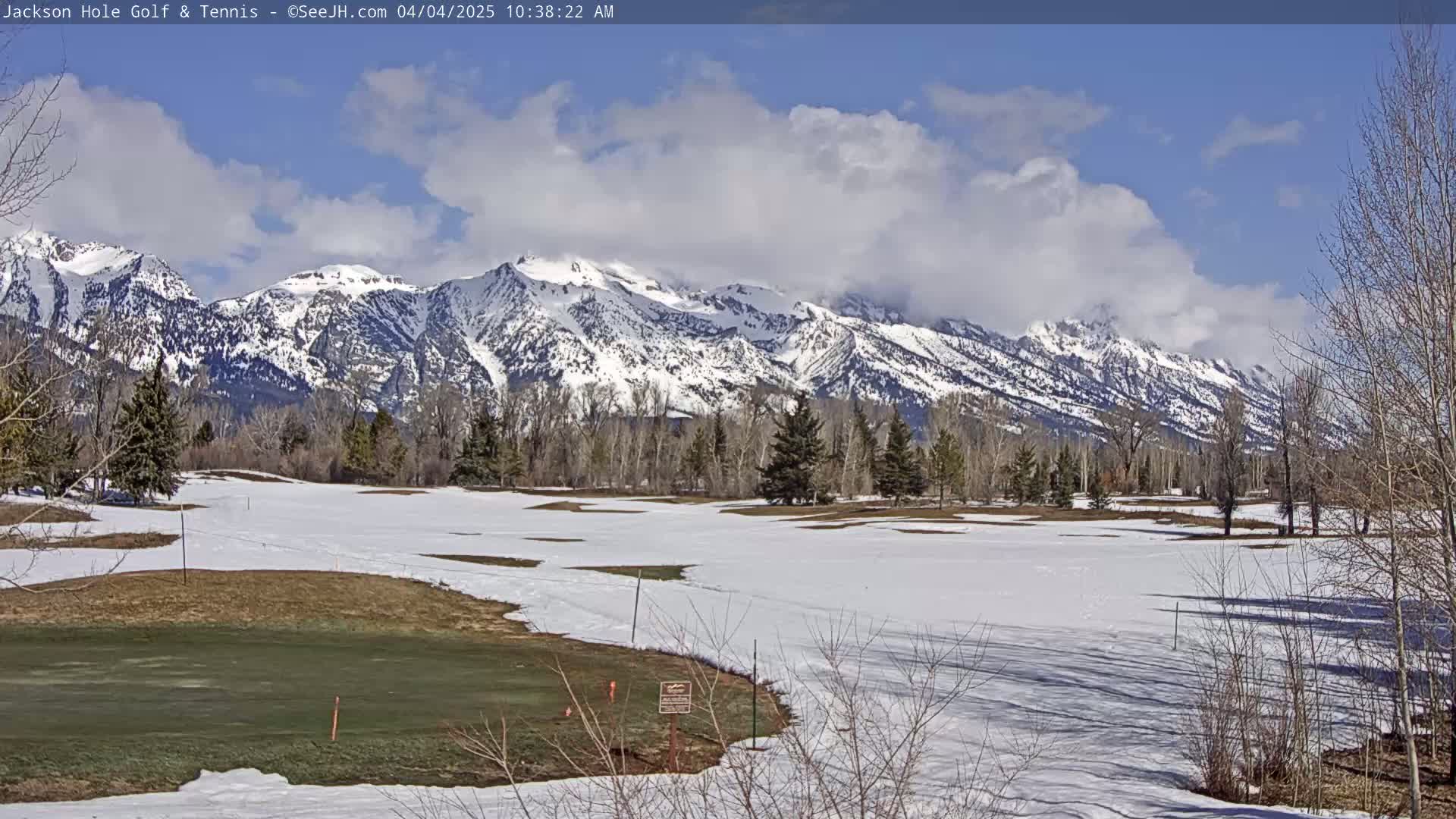 A partially snow-covered golf course stretches out before a backdrop of snow-capped mountains under a partly cloudy sky.