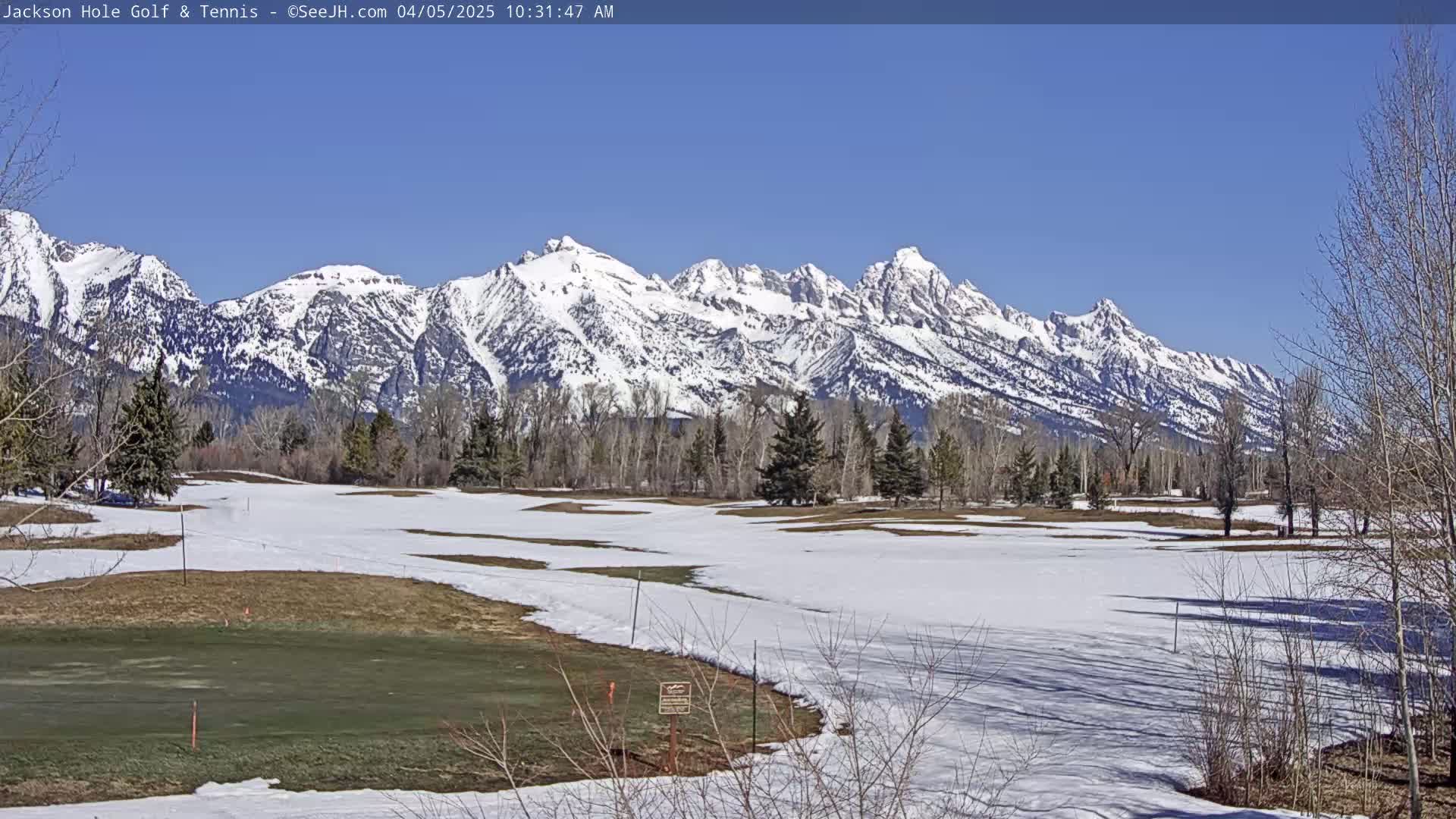 A partially snow-covered golf course stretches towards a range of snow-capped mountains under a clear blue sky.