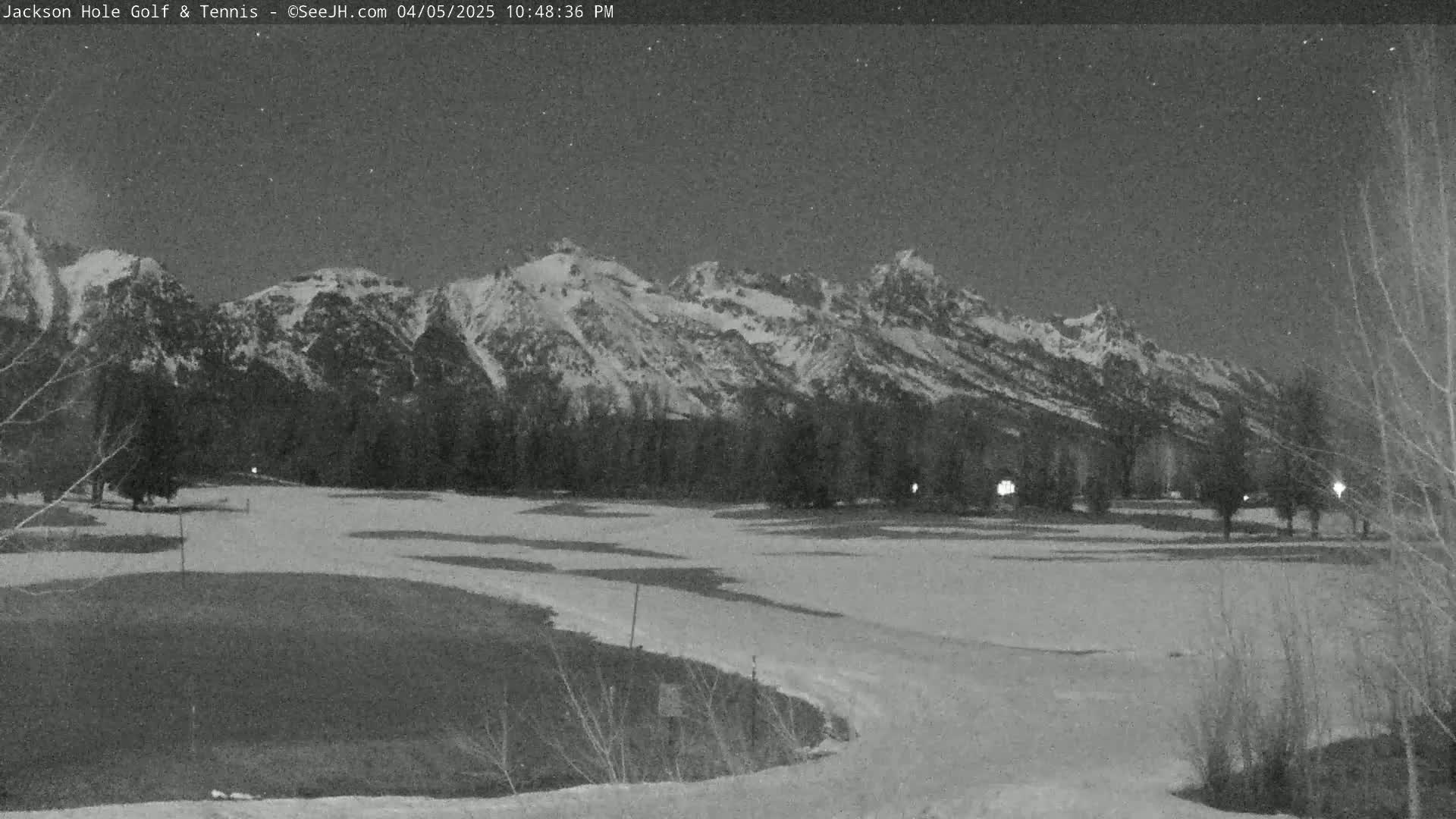 A snow-covered golf course at night is situated in front of a mountain range under a dark sky.
