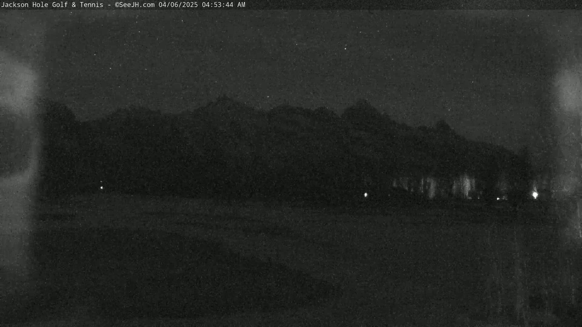 A nighttime, low-light image shows dark mountains in the distance, with a few faint lights visible near the bottom, under a clear, starry sky.