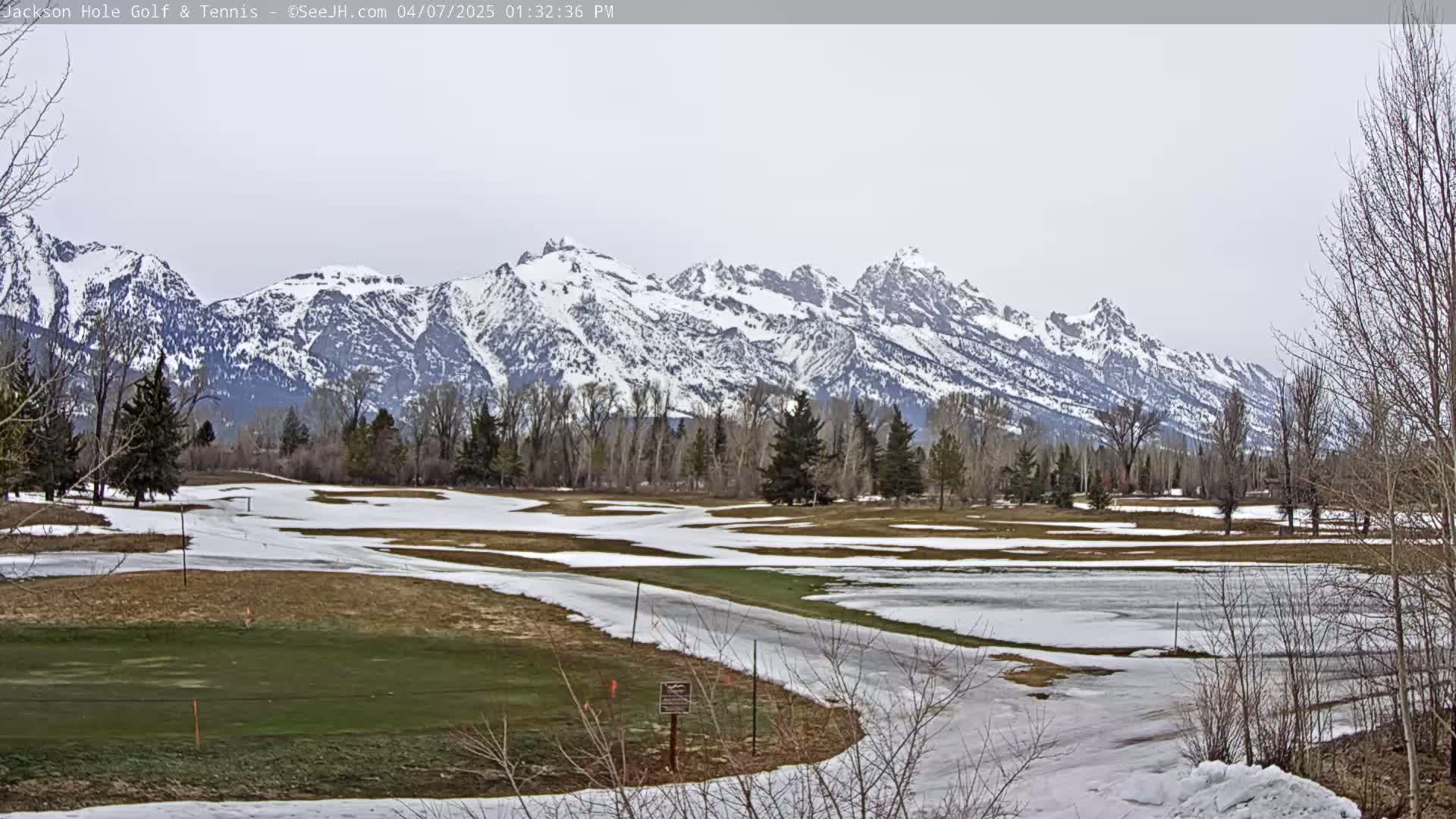 A snow-covered golf course sits in front of a range of snow-capped mountains under an overcast sky.