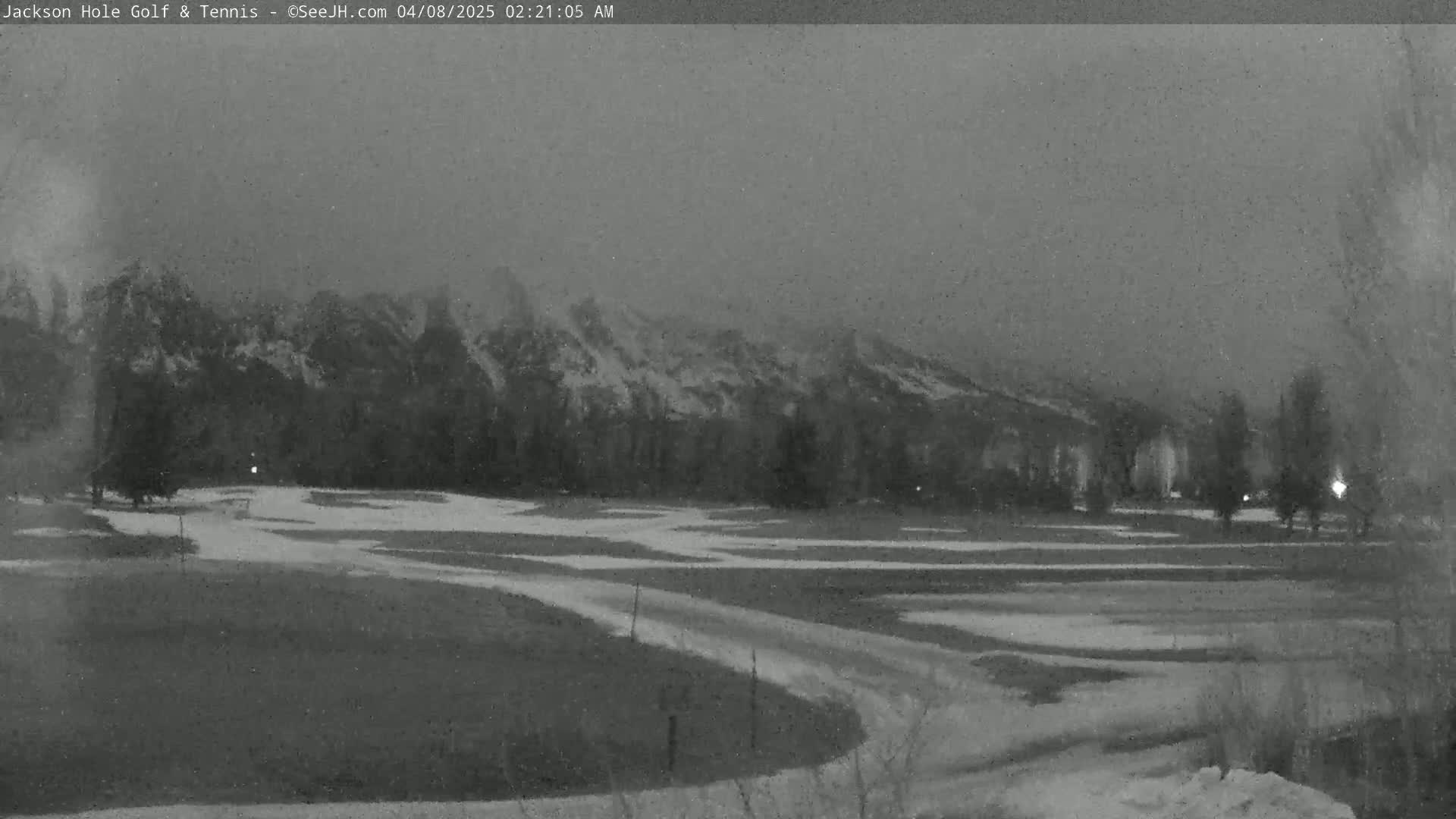 A snow-covered golf course is seen at night, with a mountain range in the background under a hazy sky.