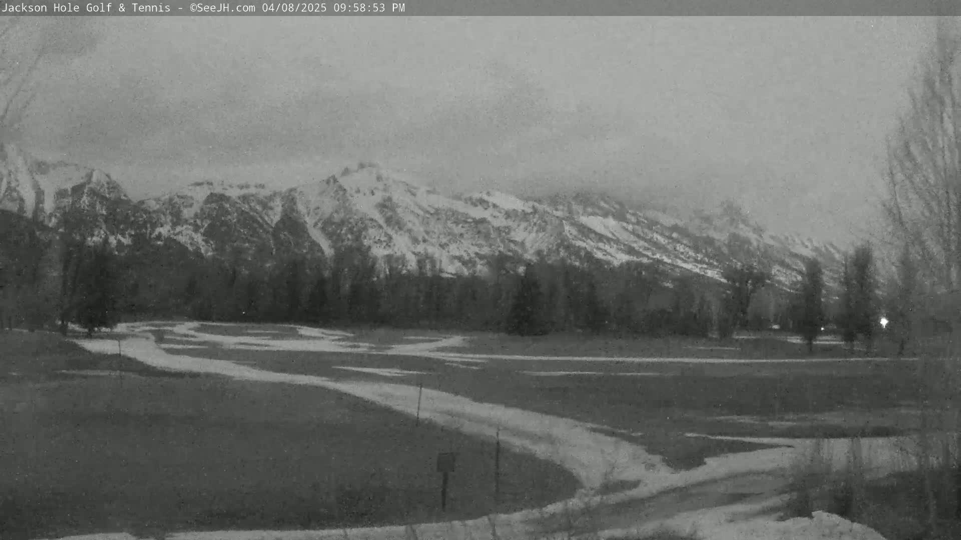 A snow-capped mountain range is visible in the distance behind a flat, snow-dusted field with winding paths, under an overcast sky.