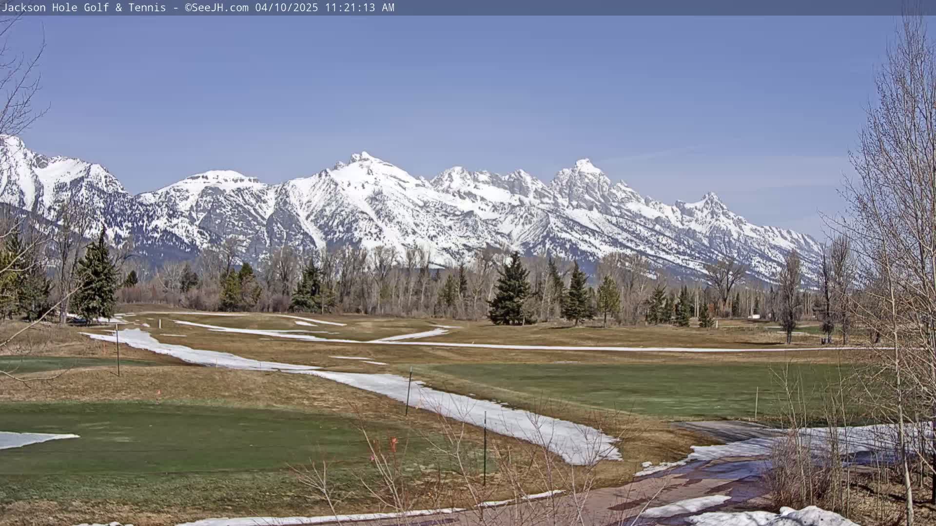 A snow-dusted golf course with patches of melting snow stretches towards a background of snow-capped mountains under a clear blue sky.