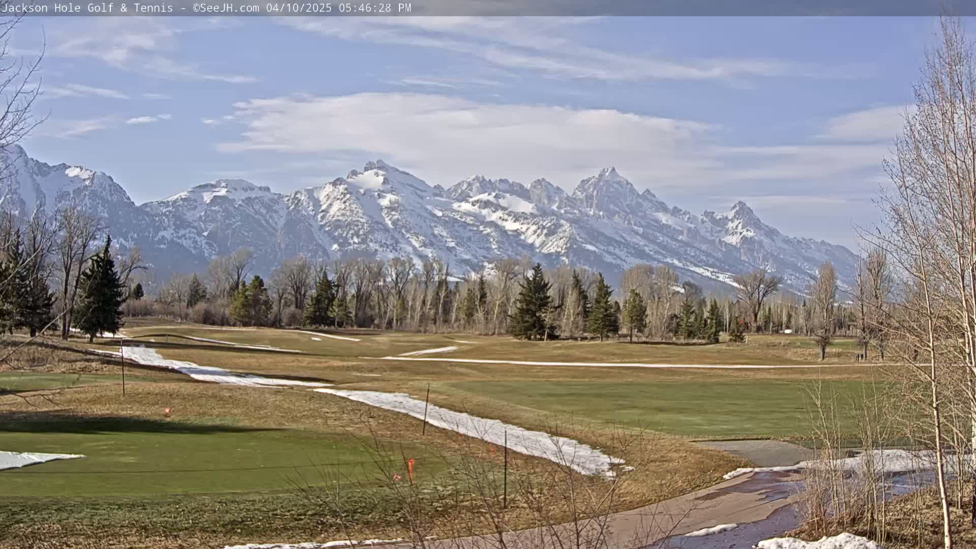 A partly sunny day reveals a snow-capped mountain range in the background of a mostly green golf course with patches of snow and bare trees.