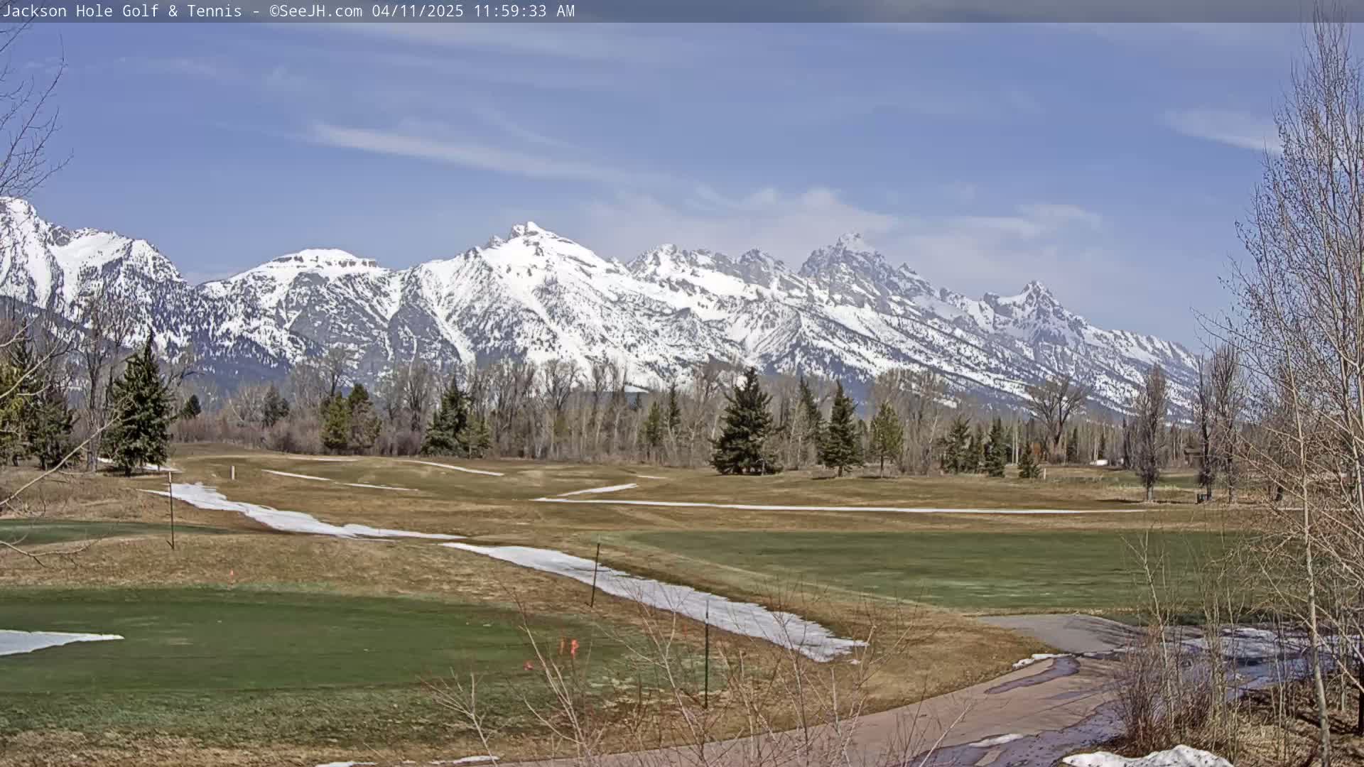 A snow-capped mountain range is visible in the background of a mostly green golf course with patches of snow and bare trees under a mostly clear, bright sky.