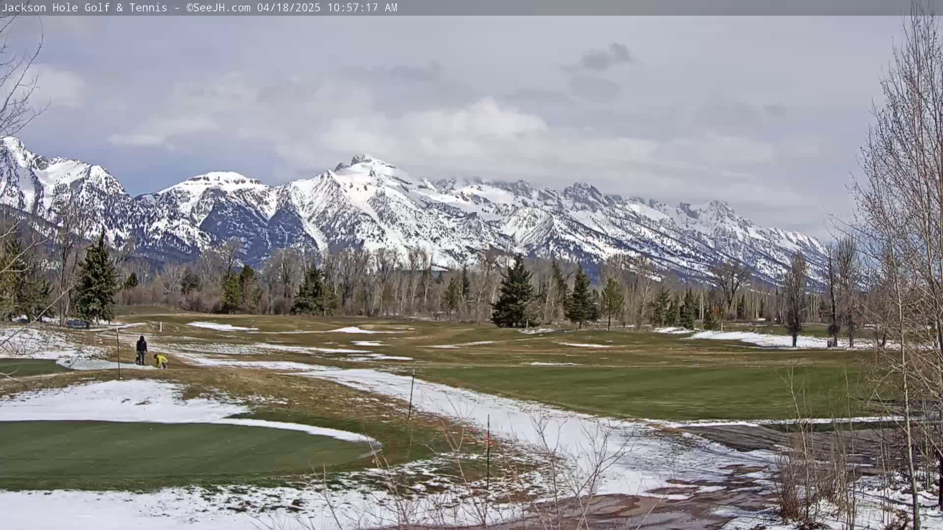 A partially snow-covered golf course stretches out before a backdrop of snow-capped mountains under a mostly cloudy sky.