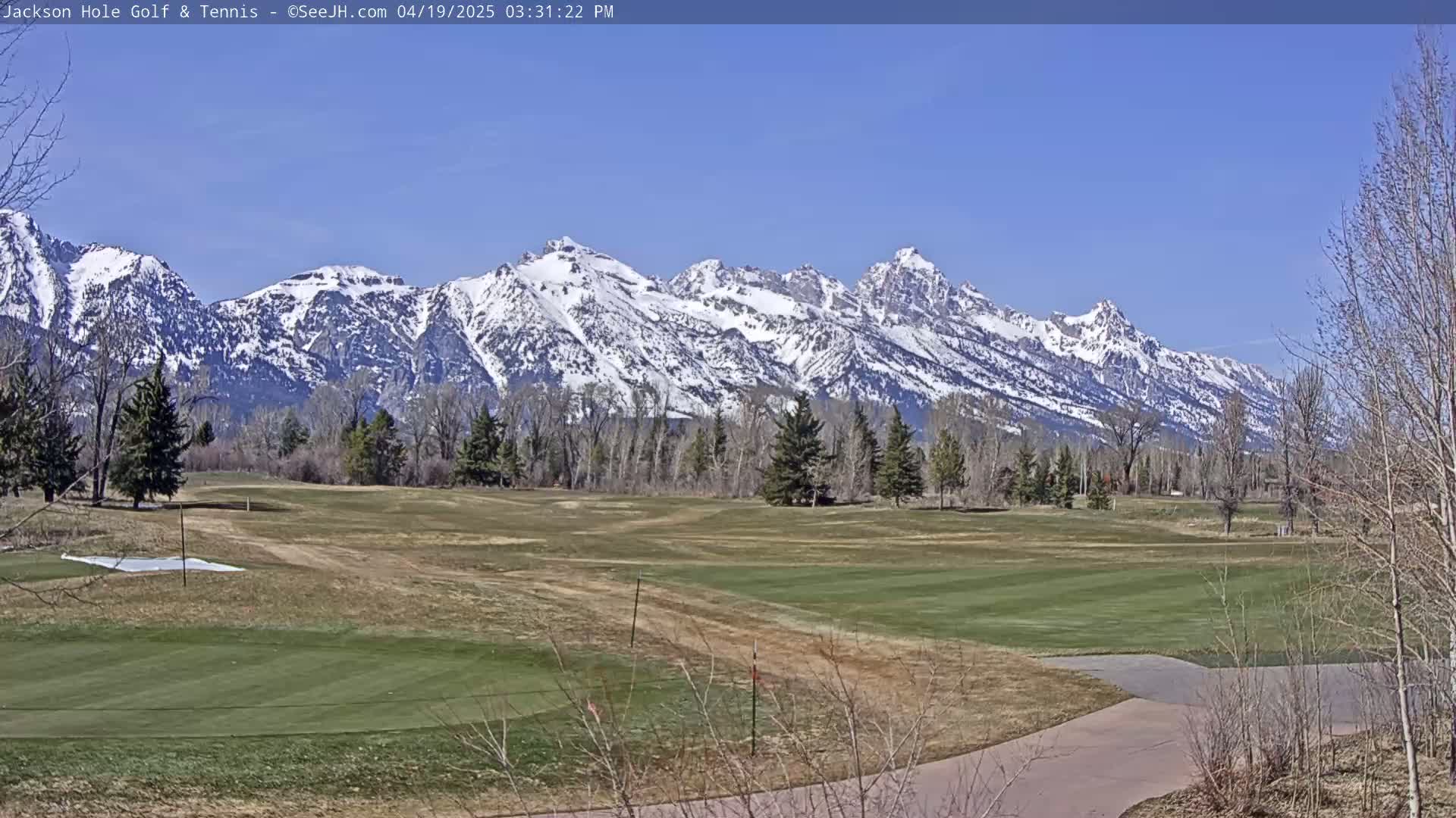 A snow-capped mountain range is visible in the background of a mostly brown and green golf course on a sunny day.