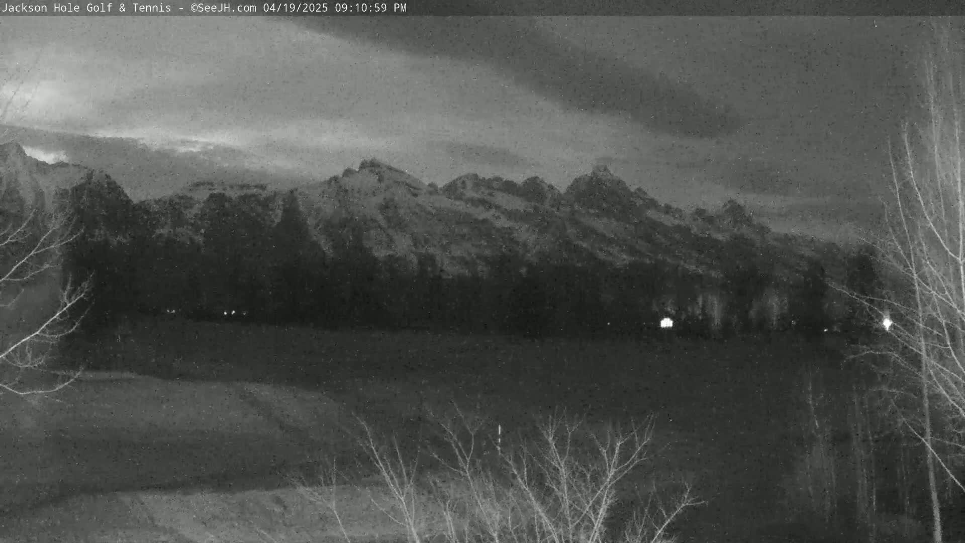 A nighttime, grayscale view shows snow-capped mountains in the distance, with bare trees and a flat, dark foreground under a cloudy sky.
