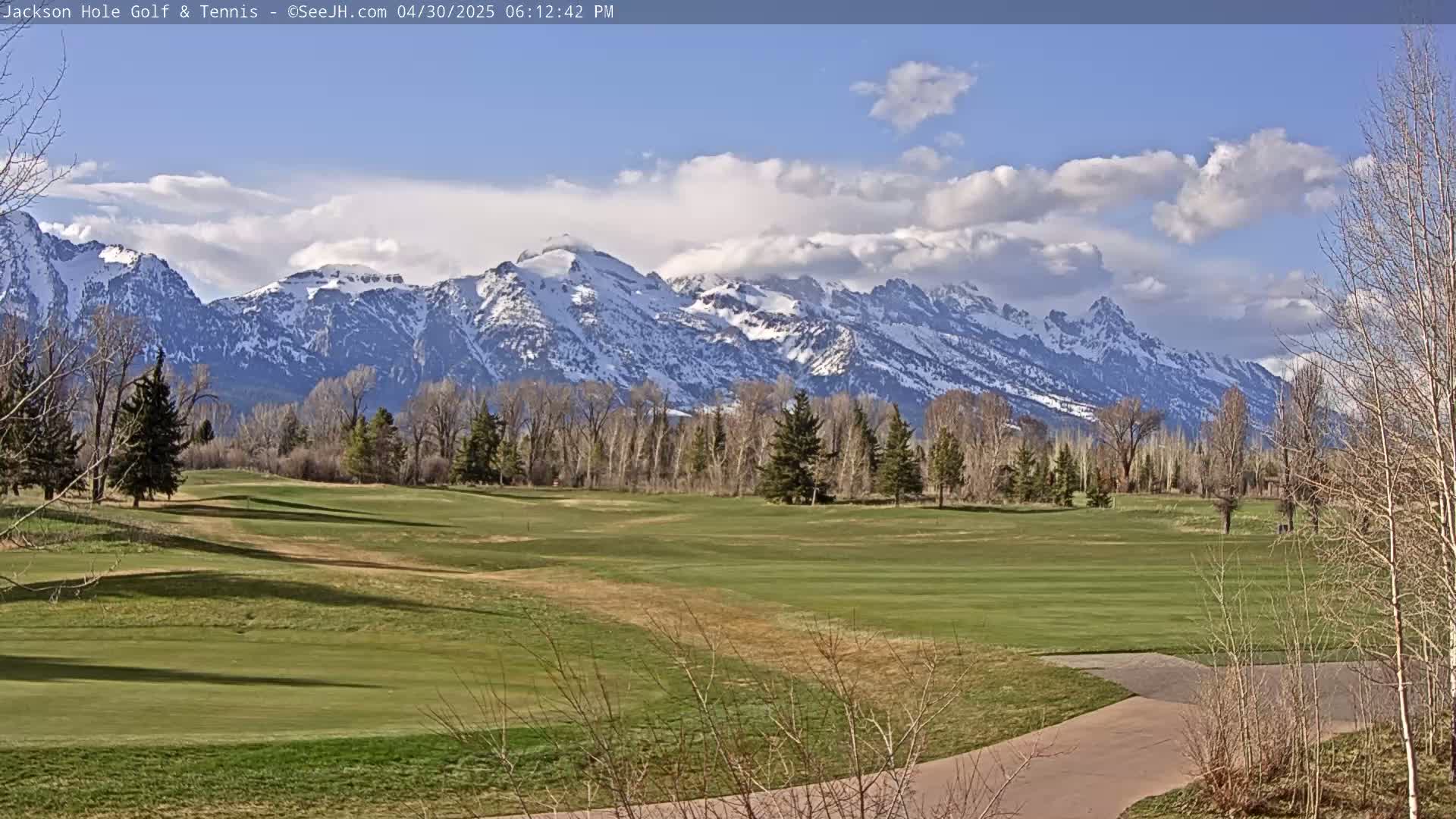 A partly cloudy day reveals a snow-capped mountain range in the background of a green golf course with leafless trees and a paved path.