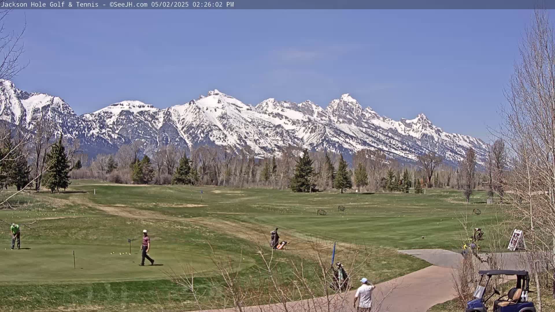 A golf course with several golfers playing, situated in front of a snow-capped mountain range under a clear, sunny sky.