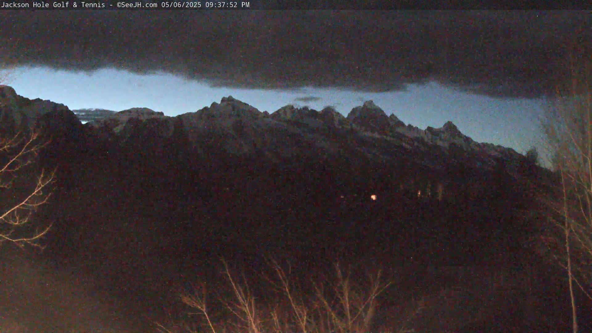 A dark, overcast night reveals snow-capped mountains in the distance, with bare trees in the foreground.