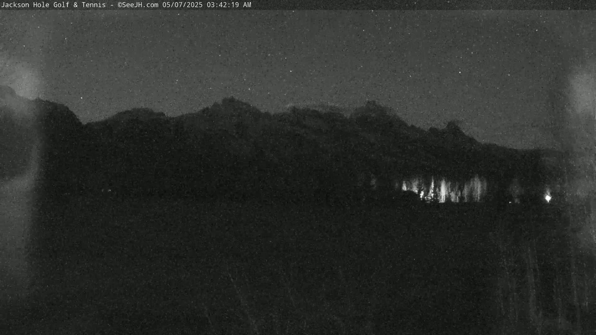 A nighttime view of dark mountains under a starry sky, with a line of lights faintly visible in the distance.